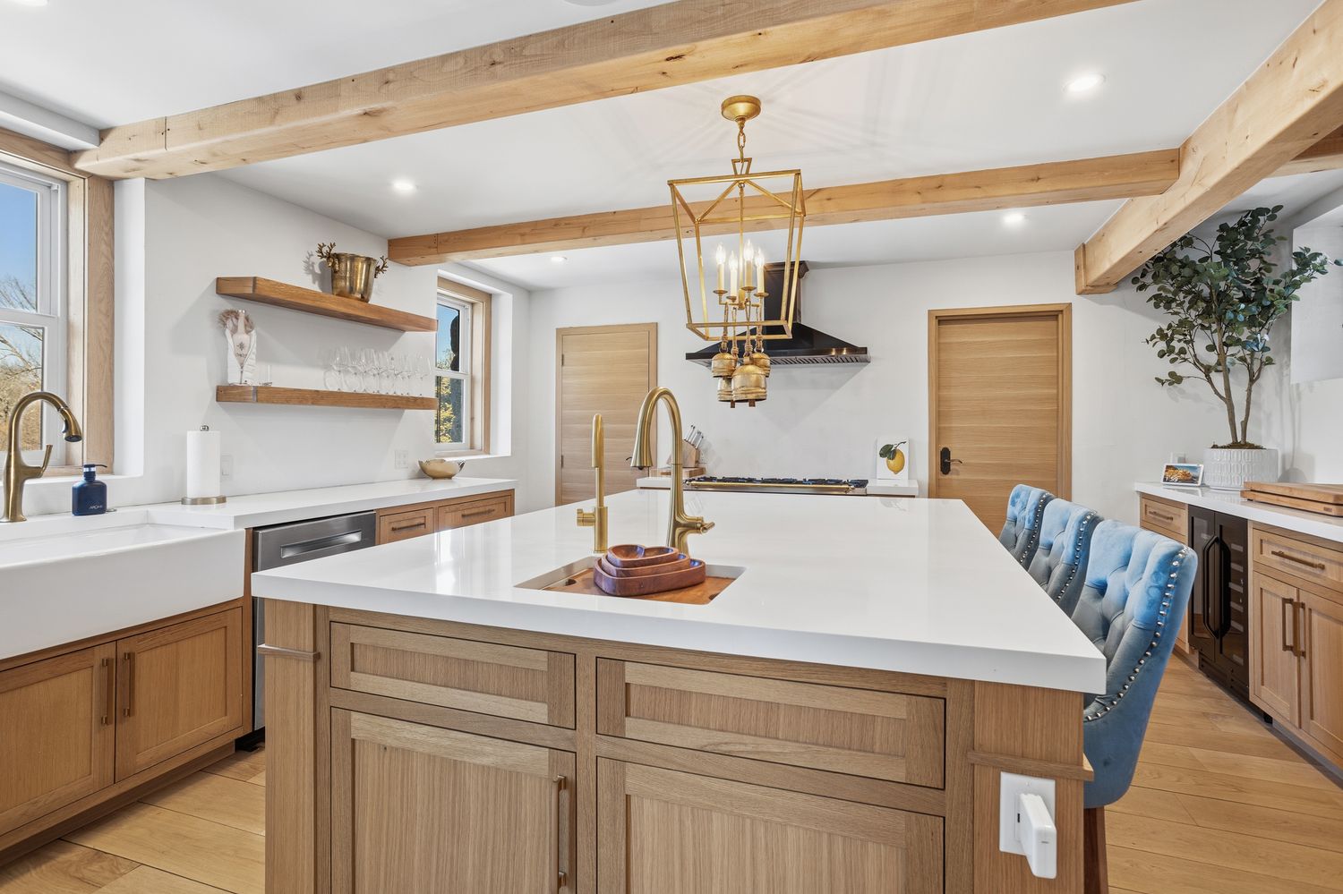 This stunning, high-end kitchen features a large central island with a white quartz countertop and a built-in sink, complemented by warm wood cabinetry and exposed ceiling beams. The space is illuminated by an elegant gold lantern-style pendant light, creating a sophisticated and inviting atmosphere. With its farmhouse-style sink, open shelving, and blue upholstered bar stools, the kitchen perfectly blends rustic charm with modern luxury.