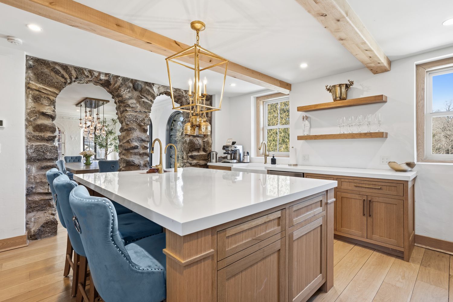 This elegant kitchen features a large central island with a white quartz countertop and light wood cabinetry, complemented by plush blue velvet bar stools. The space is defined by rustic stone archways and exposed wooden ceiling beams, creating a sophisticated blend of modern luxury and historic charm. A gold lantern-style pendant light hangs above the island, while open shelving and a windowed backsplash add to the bright, airy atmosphere.