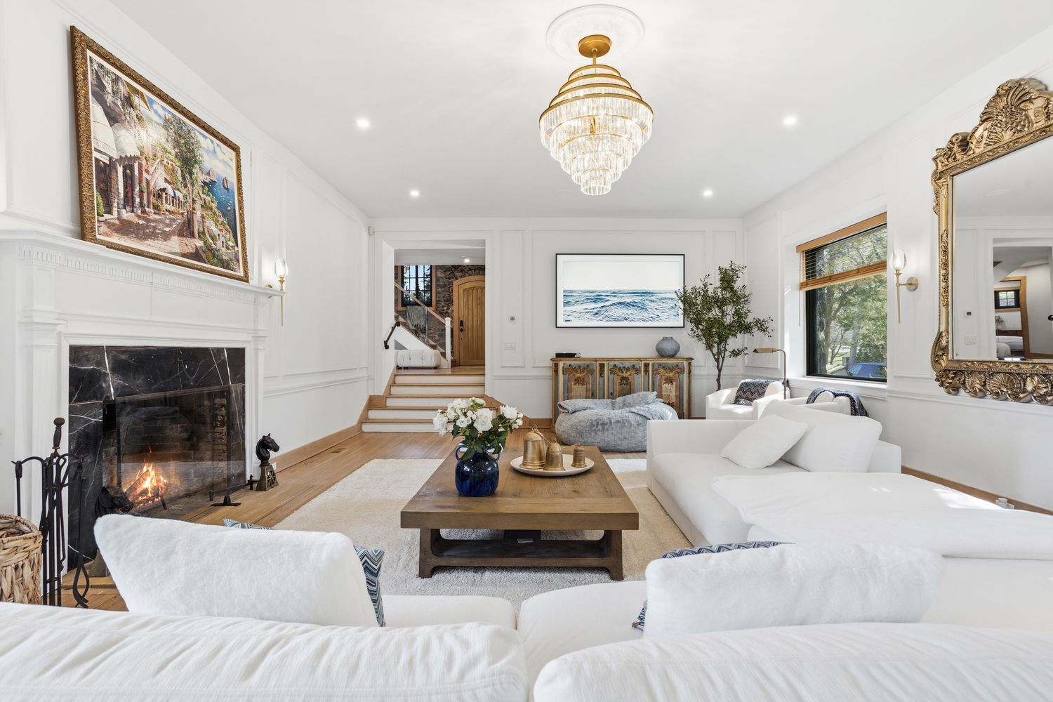 This elegant living room features a sophisticated blend of classic and contemporary design, anchored by a grand marble fireplace and a striking crystal chandelier. The space is furnished with plush white seating, a rustic wooden coffee table, and ornate gold-framed mirrors and artwork, creating a bright and luxurious atmosphere. The perspective is a wide, eye-level shot that captures the open flow of the room, including the staircase leading to an upper level.