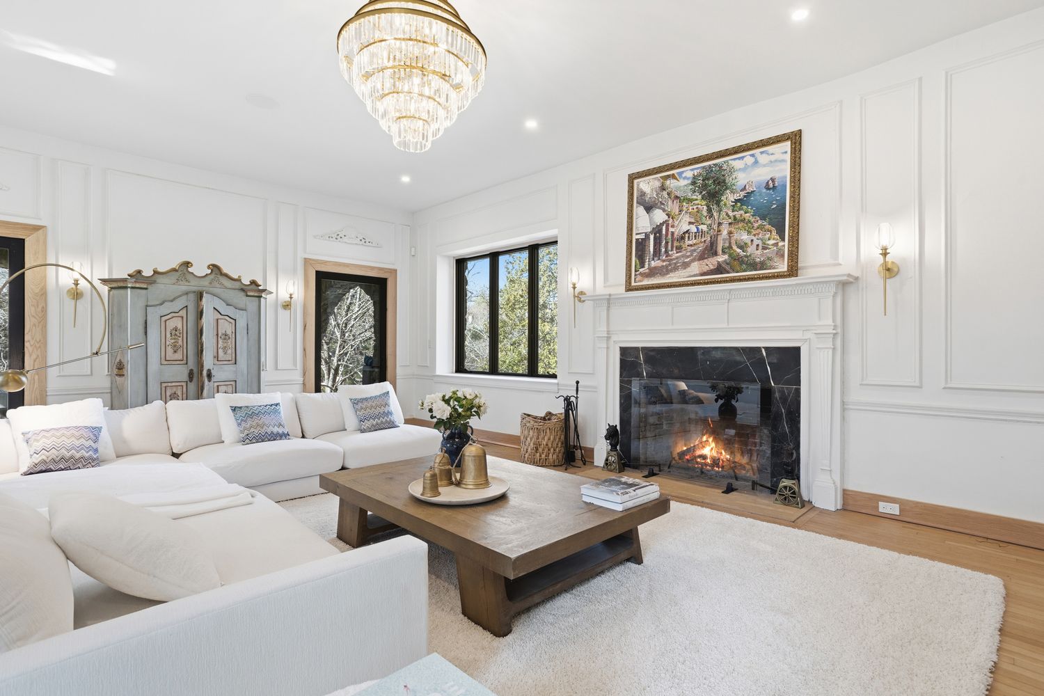 This elegant living room features a bright, airy aesthetic with white walls adorned with classic wainscoting and a large, plush white sectional sofa. A central wooden coffee table sits atop a soft white rug, facing a sophisticated black marble fireplace topped with a scenic landscape painting. The space is illuminated by a grand crystal chandelier and warm wall sconces, creating a luxurious and inviting atmosphere.