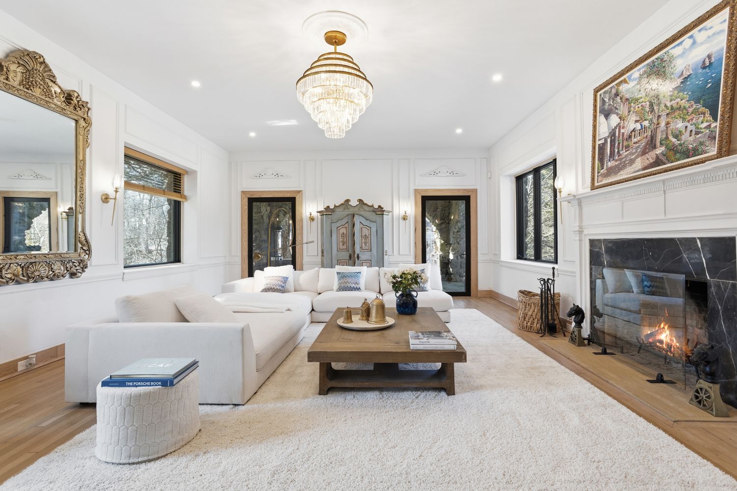 This elegant living room features a bright, airy aesthetic with white walls, wainscoting, and a plush white area rug. A large, comfortable sectional sofa faces a rustic wooden coffee table and a grand fireplace with a black marble surround, while a crystal chandelier serves as a sophisticated focal point. The space is adorned with a large ornate gold-framed mirror and a vibrant landscape painting, creating a luxurious and inviting atmosphere.