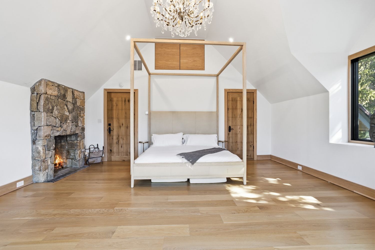 This spacious primary bedroom features a striking four-poster light wood bed frame as the central focal point, set against a backdrop of vaulted ceilings and a cozy stone fireplace. The room is bathed in natural light from a large window, highlighting the warm hardwood flooring and elegant chandelier. The overall aesthetic is a sophisticated blend of rustic charm and modern luxury, creating a serene and inviting retreat.