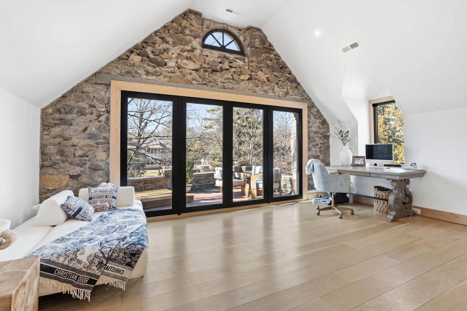 This bright and airy home office features a striking floor-to-ceiling stone accent wall and vaulted ceilings, creating a sophisticated yet rustic atmosphere. The space is anchored by a large wooden desk and a comfortable lounge area, with expansive glass doors that provide seamless access to an outdoor patio and abundant natural light. The light hardwood floors and neutral color palette enhance the room's open, tranquil, and productive feel.