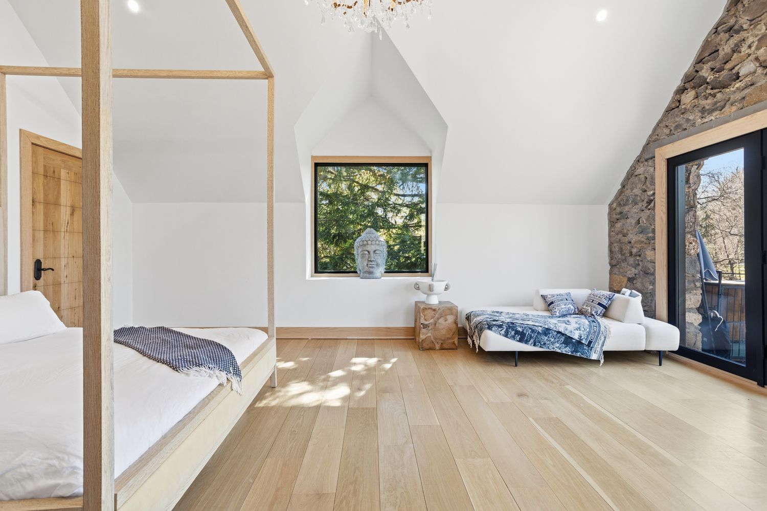 This spacious and airy primary bedroom features a minimalist aesthetic with light oak hardwood flooring and a striking four-poster wooden bed frame. The room is accented by a unique stone wall feature next to a glass door, a central window displaying a serene outdoor view with a decorative Buddha head, and a modern daybed adorned with designer textiles. The vaulted ceiling and clean white walls create a bright, tranquil atmosphere that blends rustic charm with contemporary luxury.