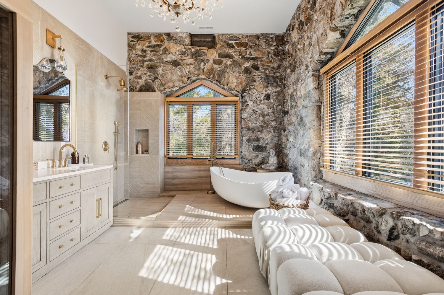 This luxurious primary bathroom features a striking blend of rustic stone walls and modern, elegant finishes. A freestanding soaking tub sits prominently on a raised platform, complemented by a sleek vanity, a glass-enclosed shower, and a plush tufted bench, all illuminated by a crystal chandelier. The room offers a serene, spa-like atmosphere with large windows that invite natural light to filter through wooden blinds.