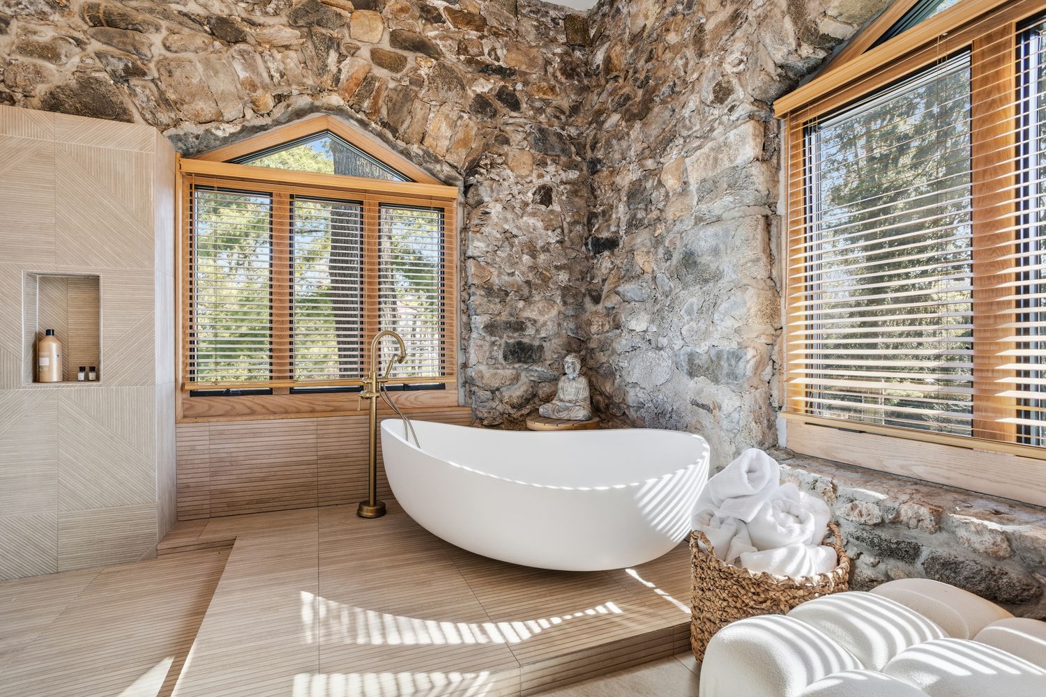 This luxurious primary bathroom features a striking blend of rustic stone walls and modern, clean-lined design elements. A sleek, white freestanding soaking tub serves as the focal point, complemented by a brass floor-mounted faucet and a serene Buddha statue nestled in the corner. Large windows with wooden blinds allow natural light to filter in, highlighting the textured tile flooring and the elegant, spa-like atmosphere.
