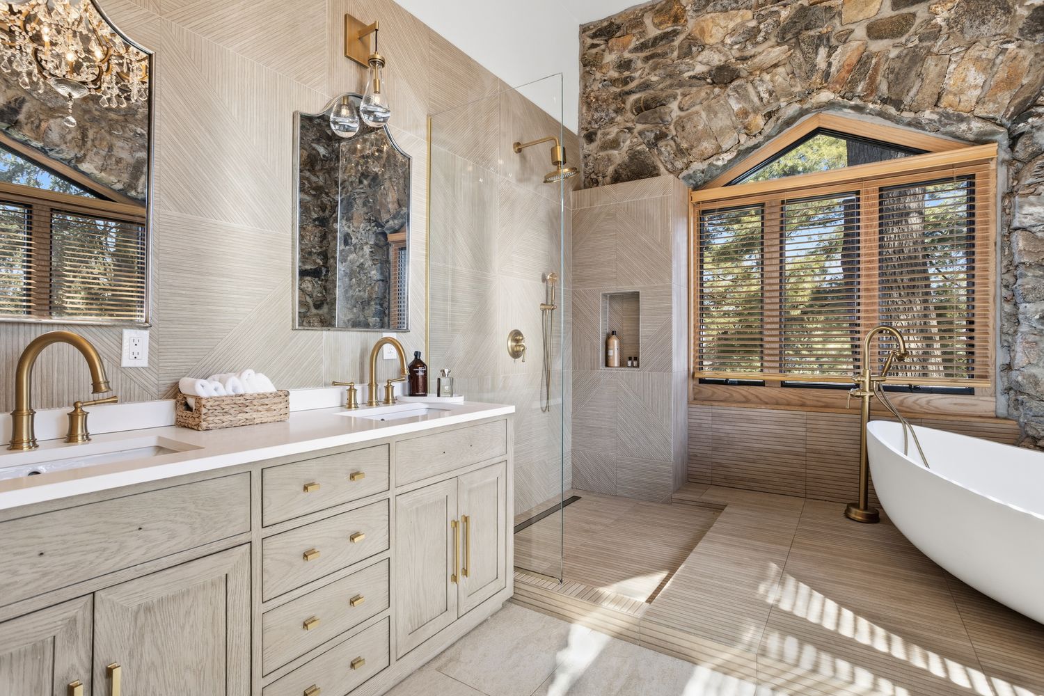 This luxurious primary bathroom features a sophisticated blend of rustic stone walls and modern, light-wood cabinetry with brass hardware. The space includes a double vanity with white countertops, a glass-enclosed walk-in shower, and a freestanding soaking tub positioned beneath a large window with wooden blinds. The combination of natural textures and elegant fixtures creates a serene, high-end spa-like atmosphere.