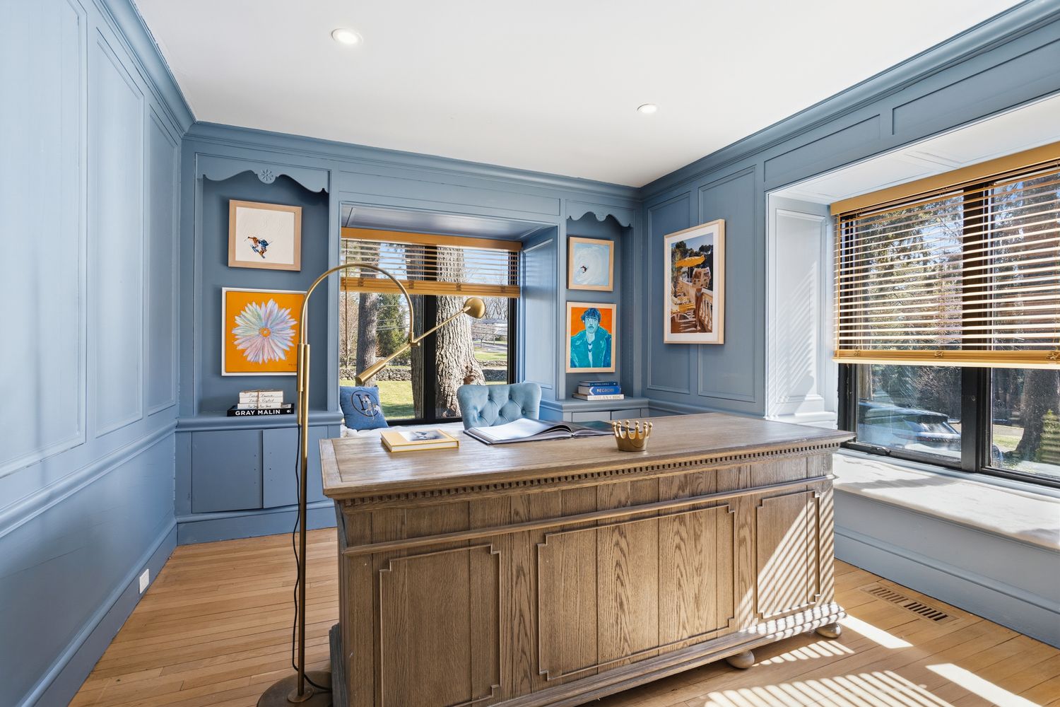 This sophisticated home office features striking blue paneled walls, a large wooden executive desk, and a comfortable blue velvet chair positioned by a window. The room is accented with vibrant framed artwork, a tall brass floor lamp, and built-in shelving, creating a professional yet stylish workspace. The perspective is a wide-angle shot that captures the symmetry of the room and the natural light streaming through the blinds.