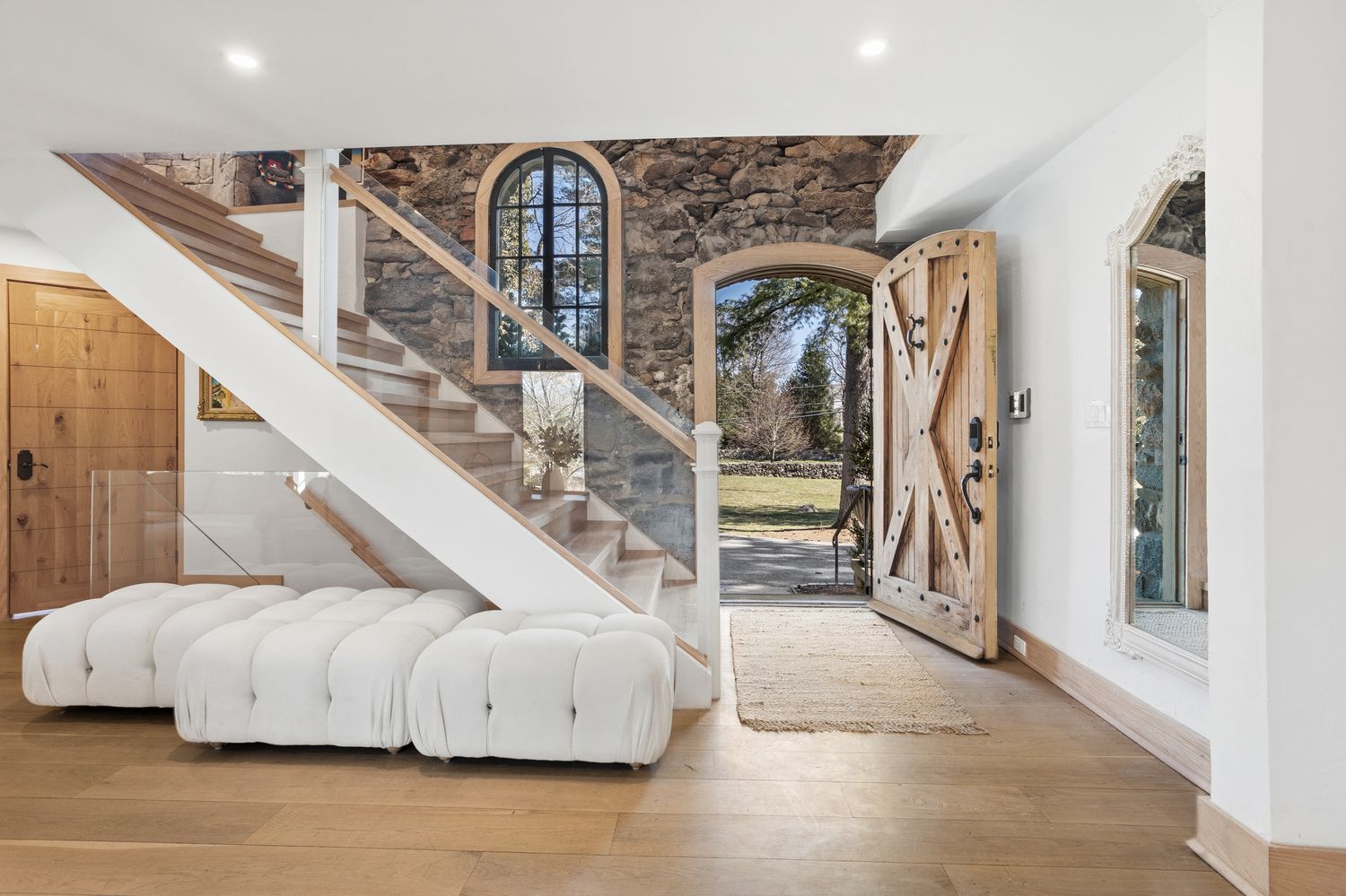 This inviting entryway features a striking blend of rustic stone walls and modern design elements, including a glass-railed staircase and light wood flooring. A plush, white tufted bench sits beneath the stairs, while a large, ornate mirror reflects the bright, open space. The heavy, wooden barn-style front door stands open, offering a welcoming view of the lush outdoor landscape.