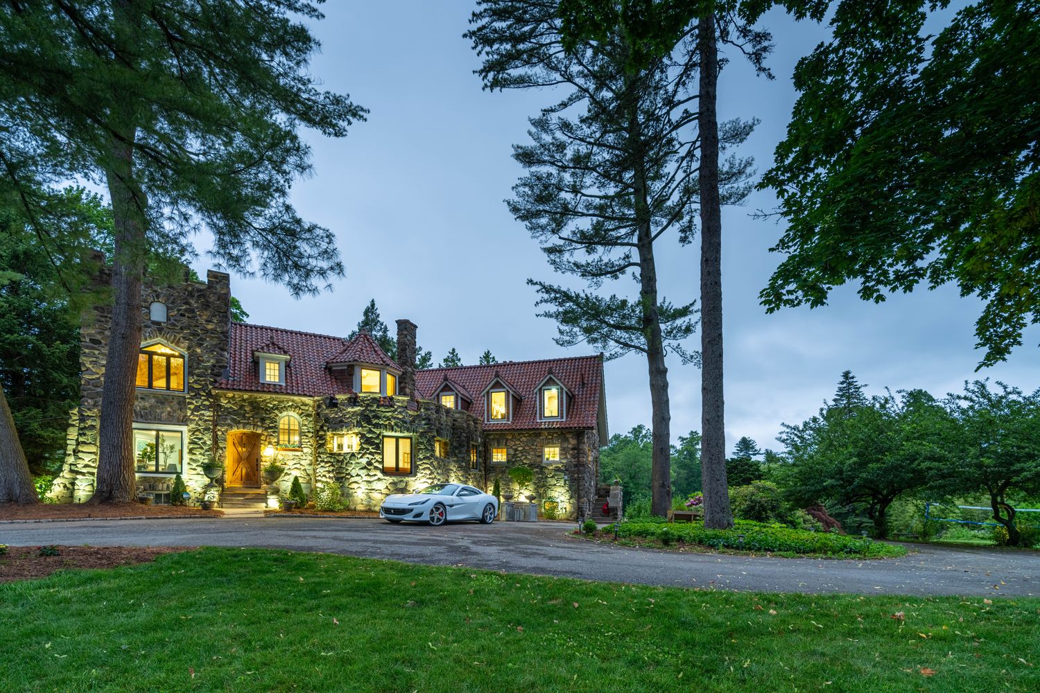 This stunning stone-clad estate features a classic European-inspired architectural style, complete with a red-tiled roof and multiple dormer windows. A sleek white sports car is parked prominently in the circular driveway, emphasizing the luxury and prestige of the property. The home is nestled among mature trees and lush greenery, creating a serene and private atmosphere at dusk.