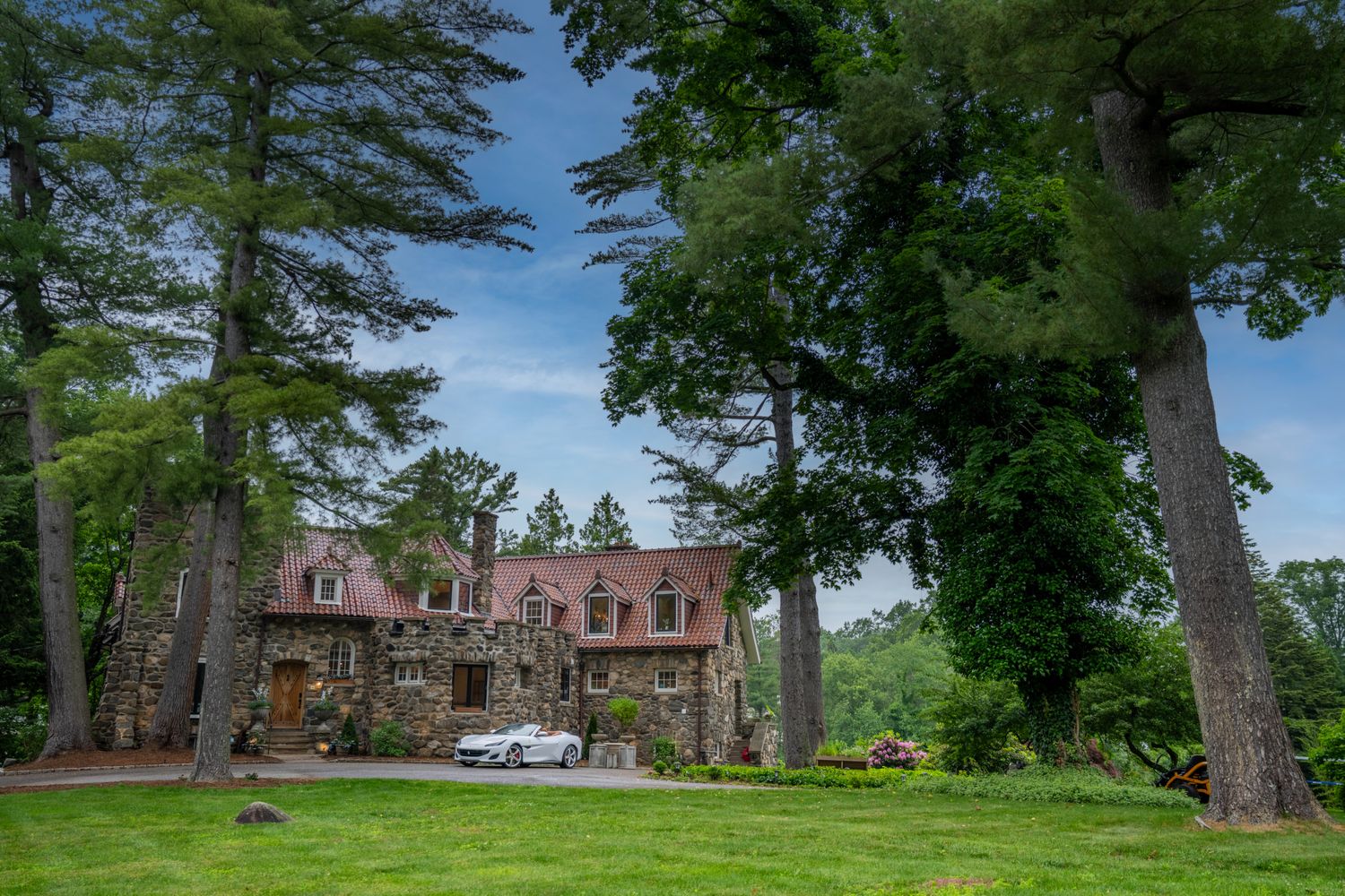 This stunning stone estate features a classic European-inspired architectural style with a prominent red-tiled roof and multiple dormer windows. A sleek white convertible is parked in the circular driveway, set against a backdrop of lush, mature trees and a sprawling green lawn. The overall impression is one of timeless elegance, luxury, and secluded privacy.