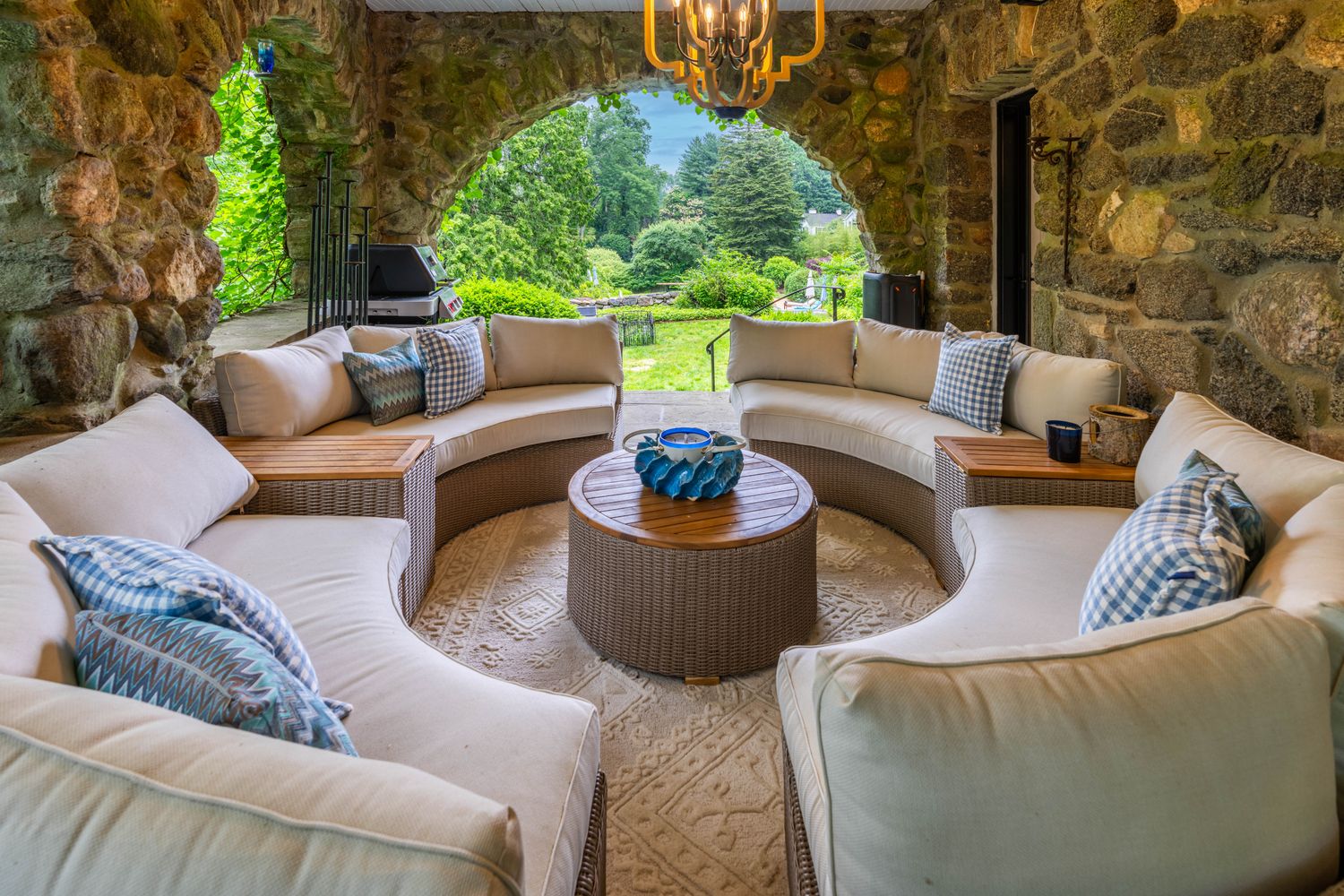 This inviting stone-walled patio features a semi-circular arrangement of plush, light-colored outdoor seating accented with blue gingham pillows. A central round wicker coffee table sits atop a textured area rug, all framed by rustic stone arches that open to a lush, verdant garden view. The space exudes a tranquil, high-end rustic charm, perfect for relaxed outdoor entertaining.