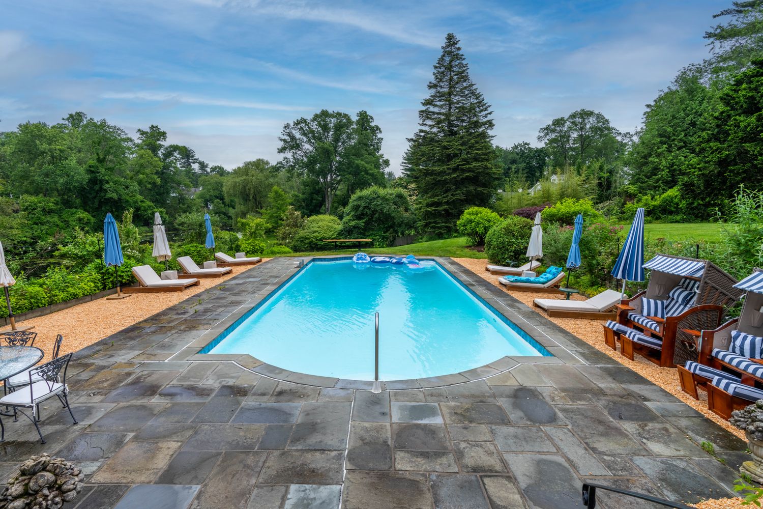 This inviting outdoor space features a rectangular swimming pool surrounded by a stone patio and gravel lounge areas. The scene is framed by lush greenery, mature trees, and multiple lounge chairs with umbrellas, creating a serene and private resort-like atmosphere. The perspective is a centered, eye-level shot looking across the pool toward the expansive backyard landscape.