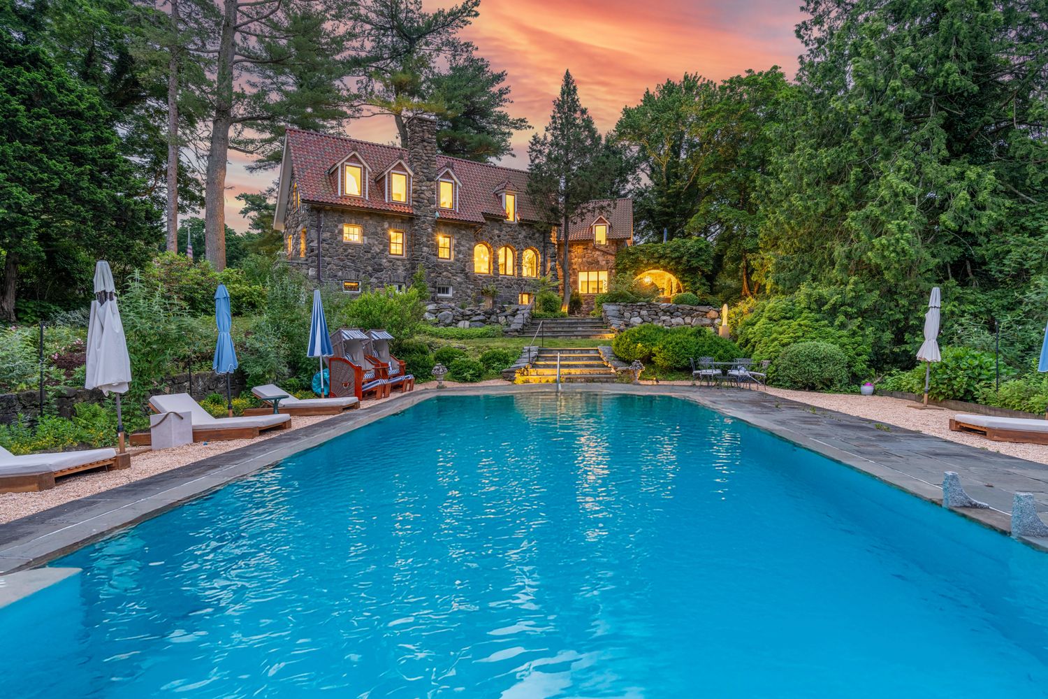 This stunning exterior shot captures a large, rectangular swimming pool set against the backdrop of a historic stone mansion at sunset. The scene features a beautifully landscaped yard with stone stairs leading up to the residence, which is illuminated by warm, inviting interior lights. The composition emphasizes a luxurious, tranquil atmosphere, perfect for outdoor entertaining and relaxation.