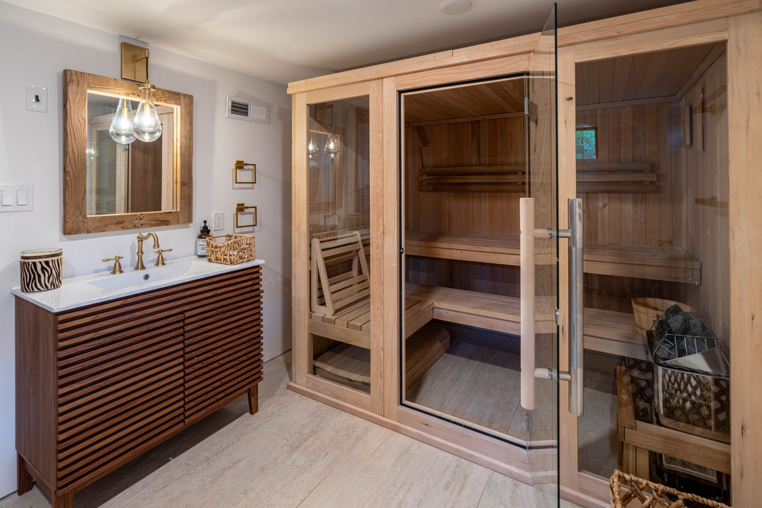 This luxurious spa-inspired bathroom features a custom-built wooden sauna with glass doors as its centerpiece. To the left, a modern vanity with a slatted wood design and a white countertop is complemented by a rustic-framed mirror and elegant gold fixtures. The space is finished with light-toned stone flooring, creating a serene and high-end atmosphere perfect for relaxation.