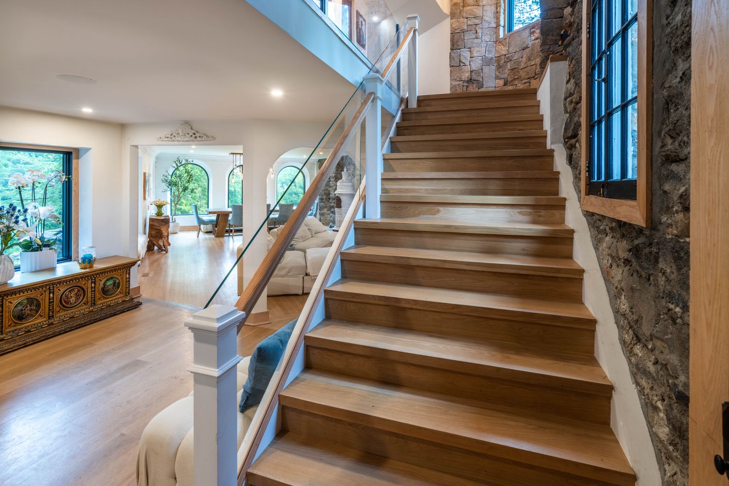This perspective captures a grand staircase with light oak treads and a modern glass railing, serving as the focal point of the home's open-concept layout. To the right, a rustic stone wall adds texture and character, while the left reveals a bright, airy living area and dining space with arched windows. The combination of natural wood, stone, and clean lines creates a sophisticated, warm, and inviting atmosphere.