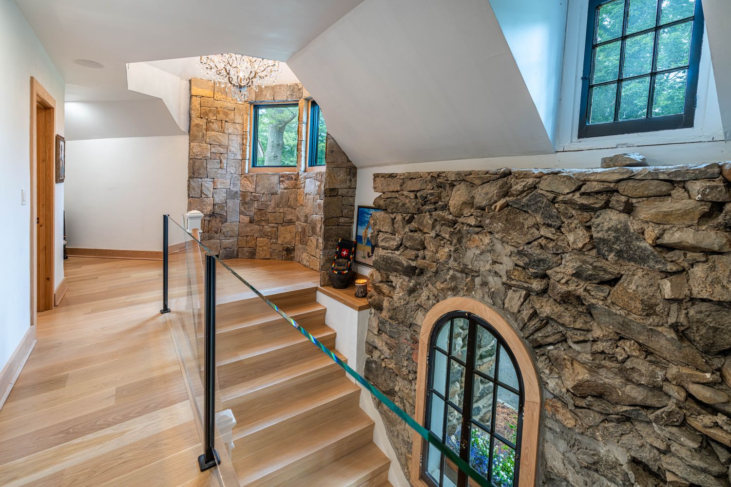 This elegant staircase landing features a striking blend of rustic stone walls and modern design elements, including a sleek glass railing and light oak hardwood flooring. A dramatic crystal chandelier hangs above the landing, while an arched window set into the stone wall provides a unique architectural focal point. The perspective captures the transition between levels, highlighting the sophisticated contrast between the rugged masonry and the clean, contemporary finishes.