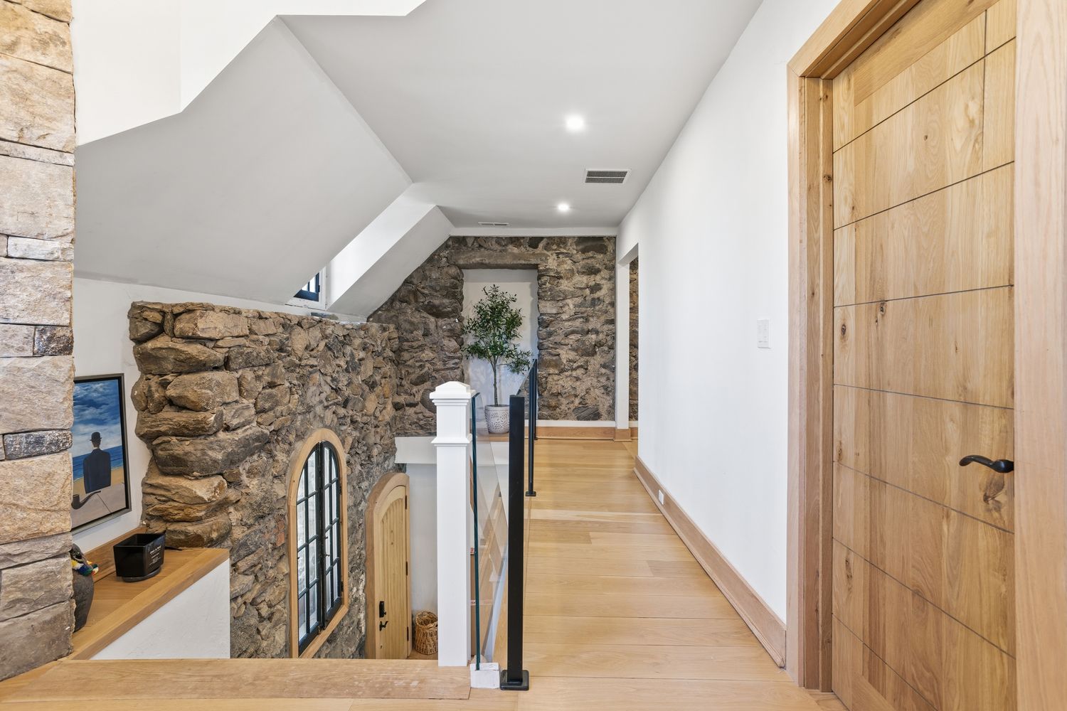 This elegant hallway features a striking contrast between rustic stone walls and modern, clean white surfaces. The space includes light-toned hardwood flooring, a contemporary wooden door, and a staircase with a sleek railing that overlooks a lower level. The architectural blend of natural textures and minimalist design creates a sophisticated and inviting atmosphere.