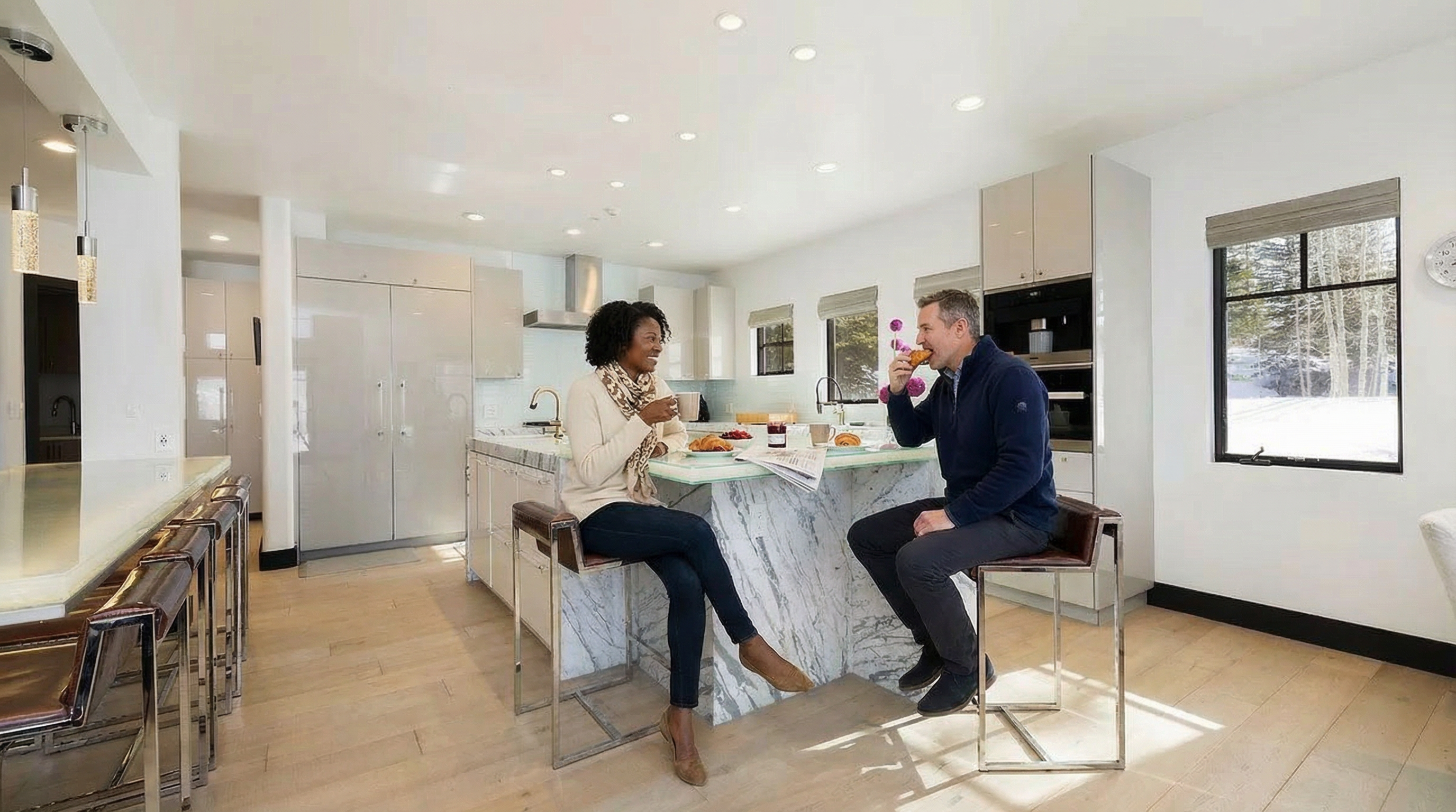 This is a bright and modern kitchen featuring a large island with marble accents. Two people are seated at the island, enjoying a meal. The kitchen has sleek, white cabinetry and stainless steel appliances, contributing to a clean and inviting aesthetic.