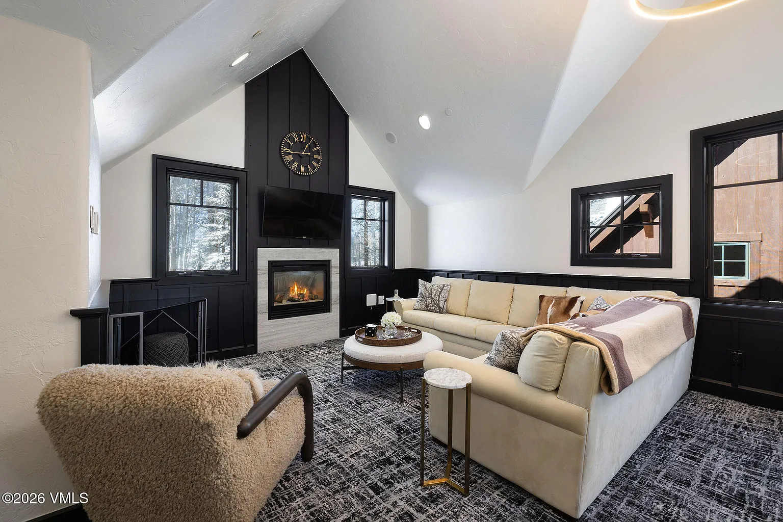 This is an interior shot of a living room featuring a fireplace, television, and comfortable seating area with sofas and an armchair. The room has an interesting architectural design with angled ceilings, with black accents on the walls and trim providing a contrast to the white walls. The carpet is a modern design with a neutral palette pulling the room together.