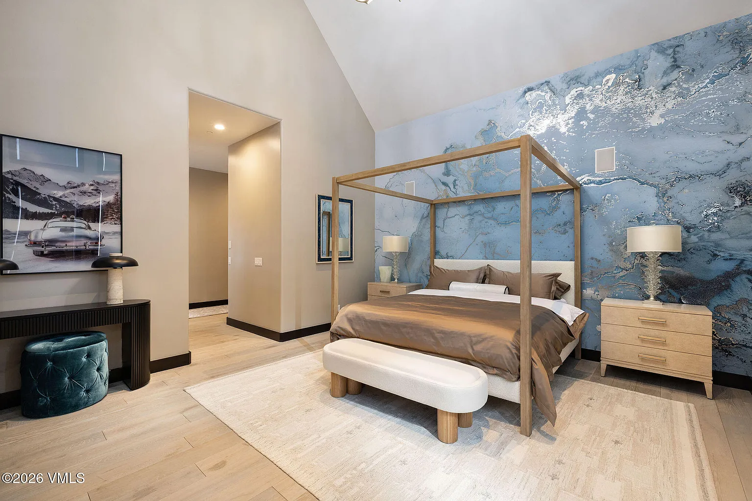 This is a primary bedroom featuring a modern four-poster bed with a light wood frame and a luxurious brown comforter. The accent wall behind the bed showcases a blue marble-like design with silver accents. The room is decorated with matching bedside tables and lamps, and a white bench sits at the foot of the bed, complemented by a plush teal ottoman and artwork nearby.