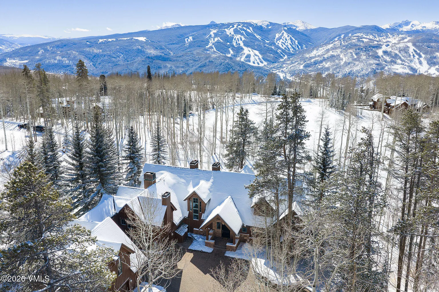 This aerial view captures a luxurious mountain home enveloped in a blanket of snow. The dark wood exterior contrasts beautifully with the snow-covered roof and surrounding landscape. The property is nestled amidst a forest of snow-dusted trees, with snow-covered mountains visible in the background offering a picturesque and serene setting.