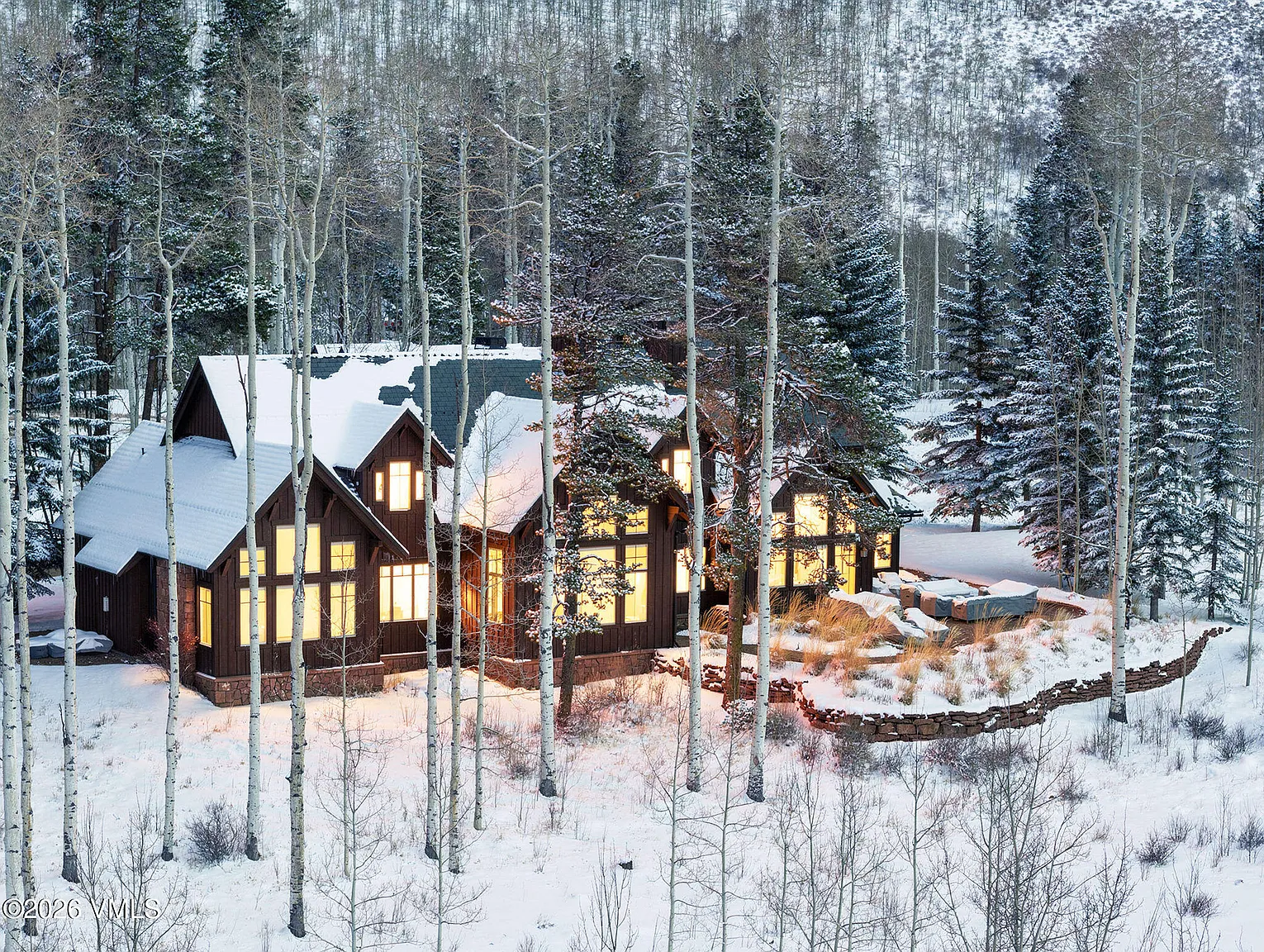 This is an eye-level exterior front view of a luxury residence in a snowy, wooded setting. The architecture features dark wood siding, steep gables, and visible windows that emit a warm, inviting glow. The landscape is heavily wooded with snow-covered aspens and evergreens, conveying a sense of seclusion and mountain elegance.