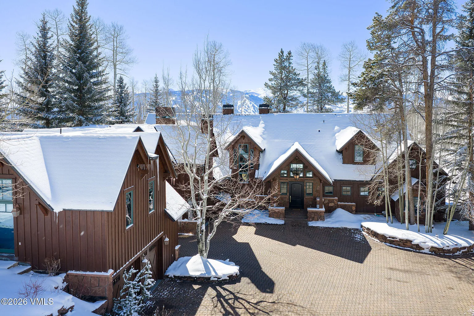 This shot captures the stately front of a mountain home in winter. The exterior features a mix of wood siding and stone accents, complemented by snow-covered roofs. The paved driveway adds to the curb appeal, and mature trees surround the property, creating a sense of privacy.