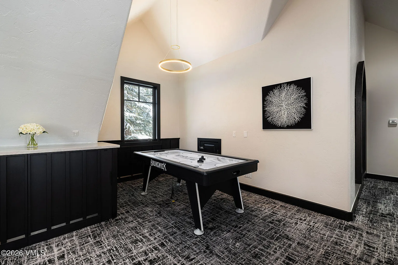 This interior shot showcases a game room featuring an air hockey table, a modern circular light fixture, and an abstract piece of artwork on the wall. A built-in black cabinet sits to the left of the window, complementing the black trim and flooring, which create a cohesive design. The space appears functional, well-lit, and designed for entertainment.