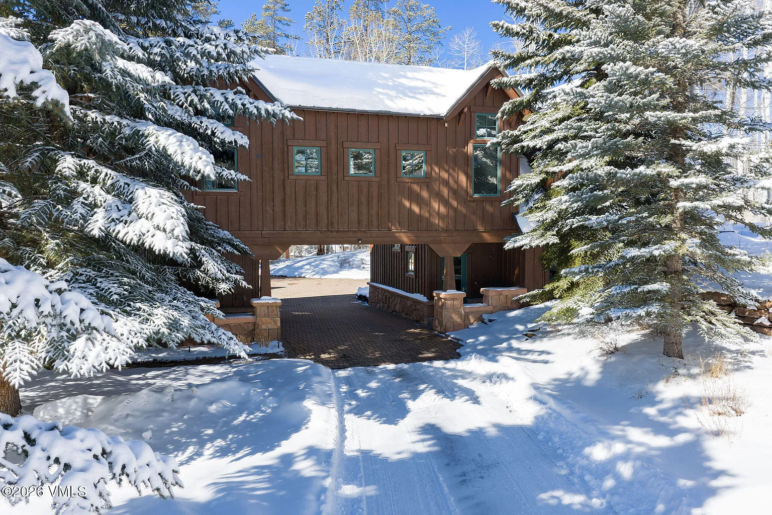 This is a front view of a unique home featuring a covered driveway connecting two portions of the house. The house has a rustic, woodsy vibe with snow-covered roof and surrounding pine trees, projecting a cozy winter retreat. The paved driveway leads directly into the covered area, offering a practical shelter for vehicles.