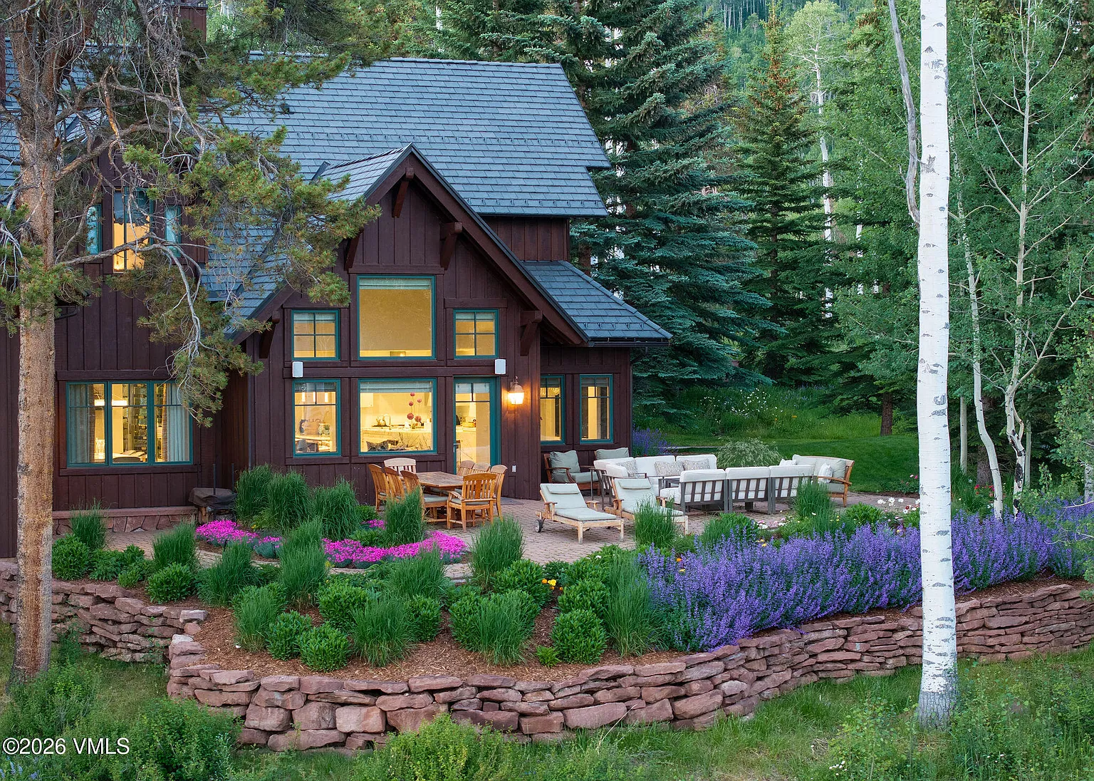 This image showcases the rear exterior of a luxurious mountain home. A stone patio extends from the house and features outdoor seating, including a dining table and lounge area. The space is beautifully landscaped with colorful flowers and shrubs, creating a serene outdoor living environment.