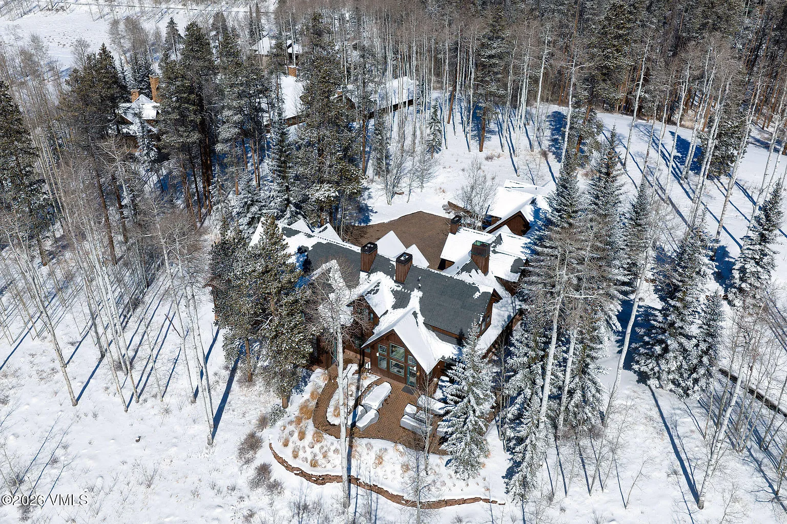 This aerial view showcases a luxury residence nestled amidst a snowy, wooded landscape. The house features a multi-gabled roof, multiple chimneys, and a stone patio area surrounded by mature evergreen trees and bare deciduous trees. The setting suggests a secluded and upscale mountain retreat.