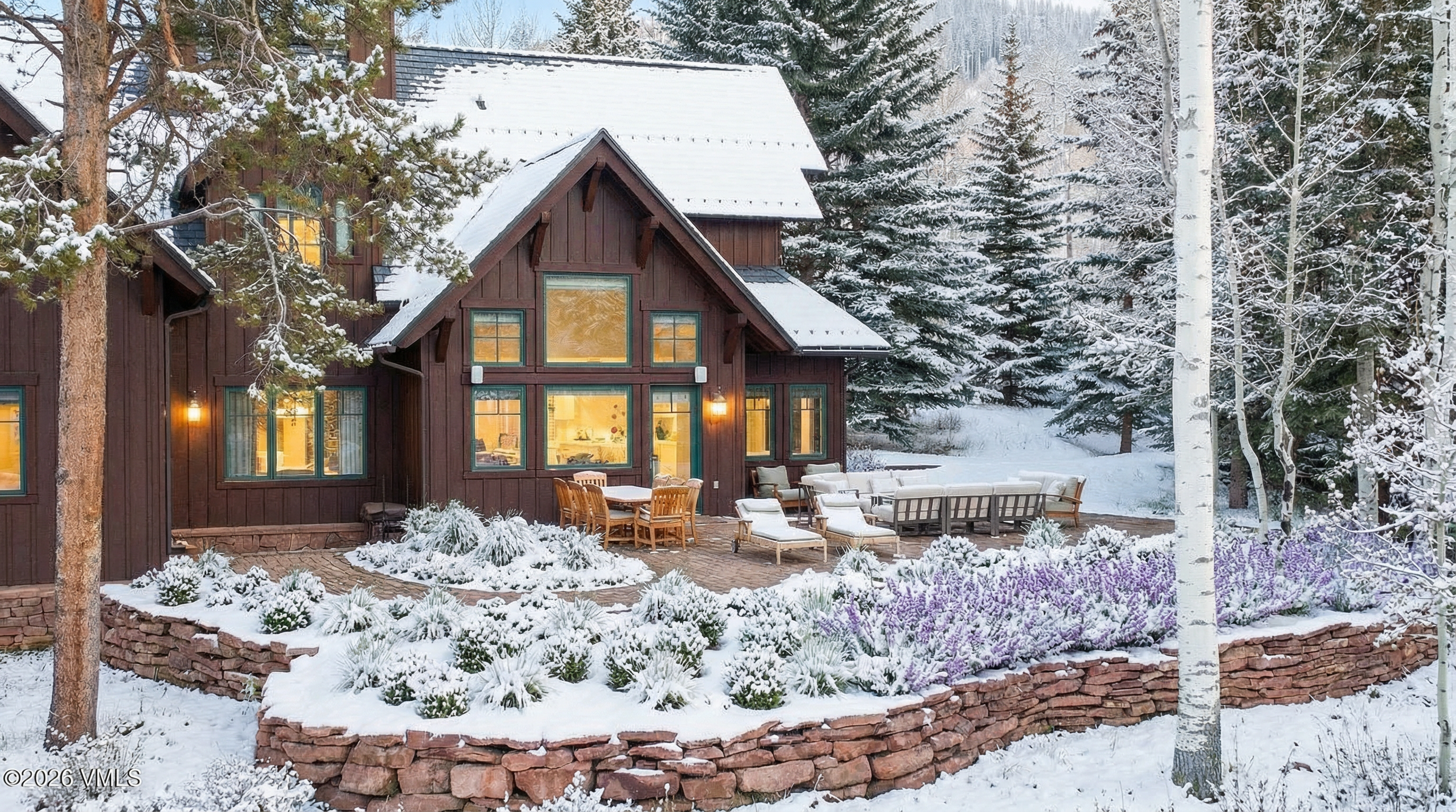 The image showcases the rear exterior of a beautiful mountain home in winter. A stone patio with outdoor furniture is visible, surrounded by snow-covered landscaping. The home features dark wood siding, green-trimmed windows emitting warm light, and a snow-covered roof, creating a cozy and inviting atmosphere.
