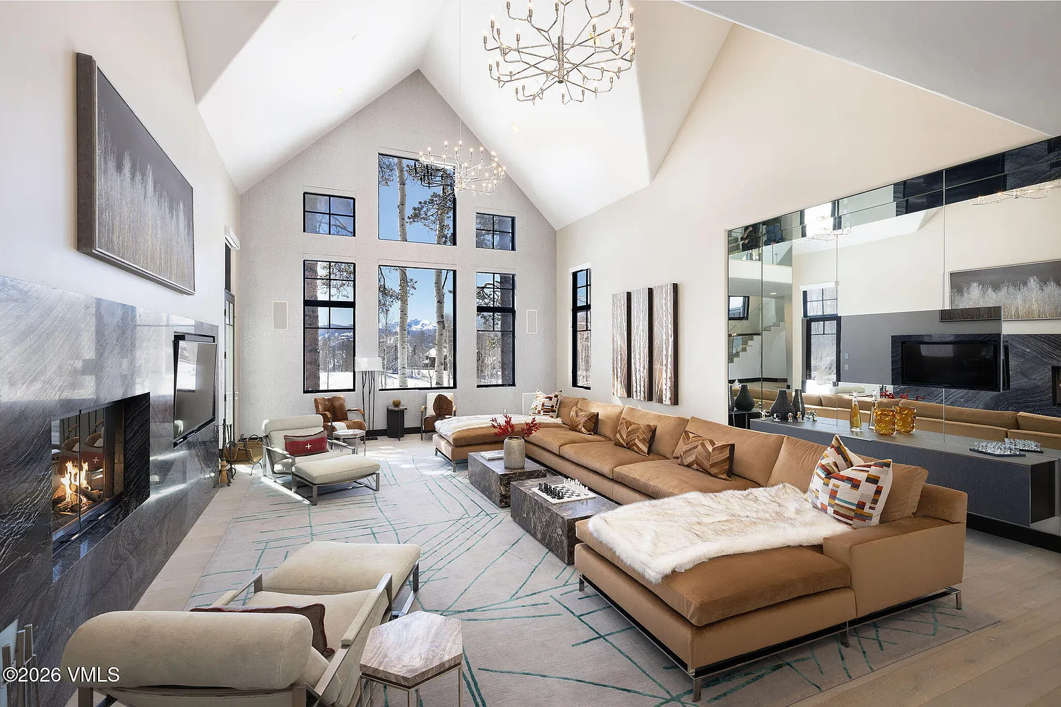 This is a bright and spacious living room featuring high vaulted ceilings and large windows that provide ample natural light and scenic views. The room includes a large sectional sofa, stylish coffee tables, and a modern fireplace, creating a comfortable and luxurious atmosphere. The design is elegant and contemporary, perfect for entertaining or relaxing.