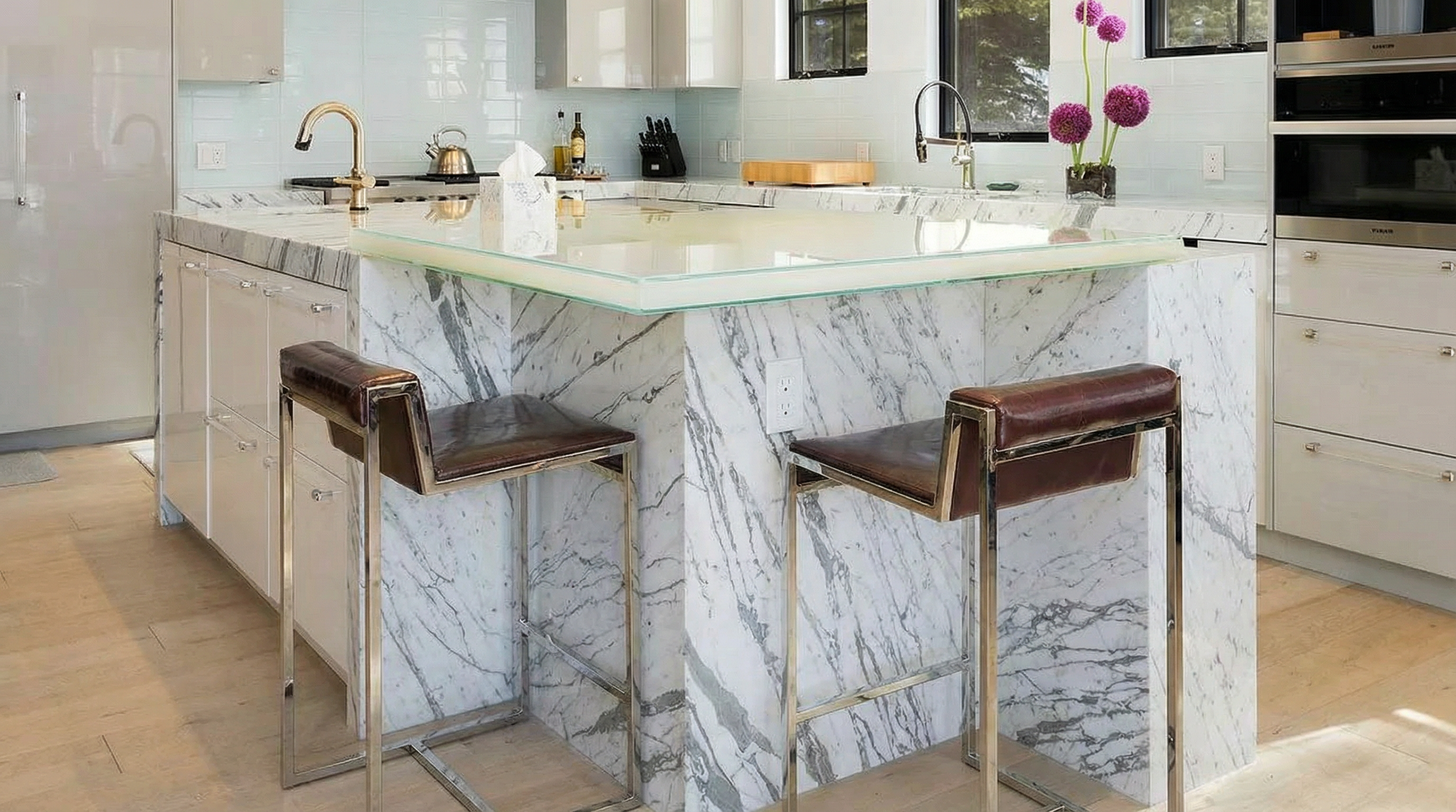 This kitchen boasts a modern design with a striking marble-clad island topped with a glass countertop extension and seating. White cabinetry and stainless steel appliances convey a clean and upscale aesthetic. The kitchen is bright and airy, indicative of a high-end property.