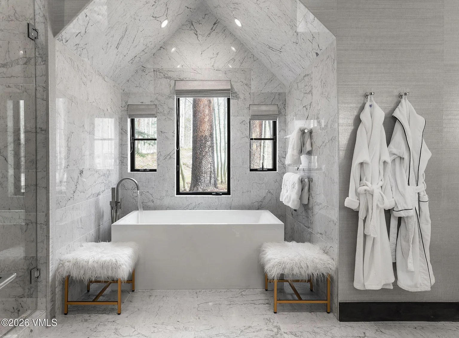 This is a luxurious primary bathroom featuring marble tiling on the walls and floor. It includes a freestanding bathtub positioned beneath a large window that offers an outdoors view. Additional features include fluffy bench seating near the tub and two white bathrobes hung on the wall, suggesting a high-end, spa-like atmosphere.