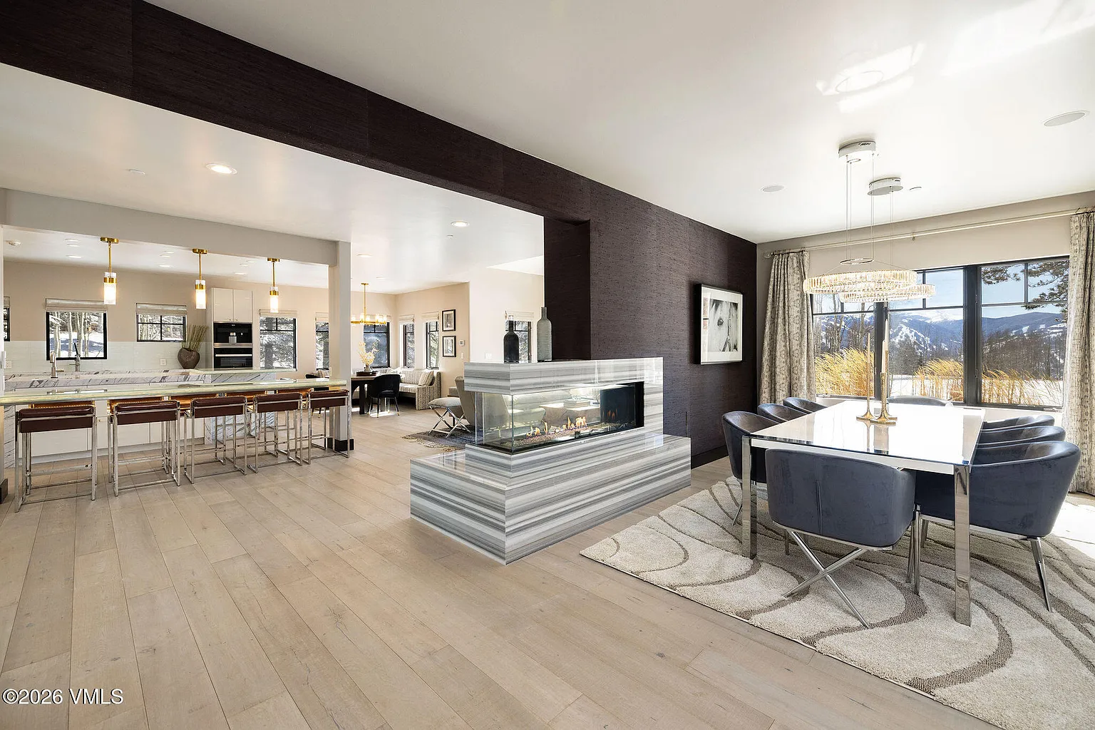 The dining room presents a contemporary setting with a sleek, white dining table surrounded by plush blue velvet chairs. A modern chandelier hangs above the table, complementing the natural light streaming through a large window with mountain views. The room is adorned with a unique marble fireplace, adding a touch of elegance and warmth to the space.