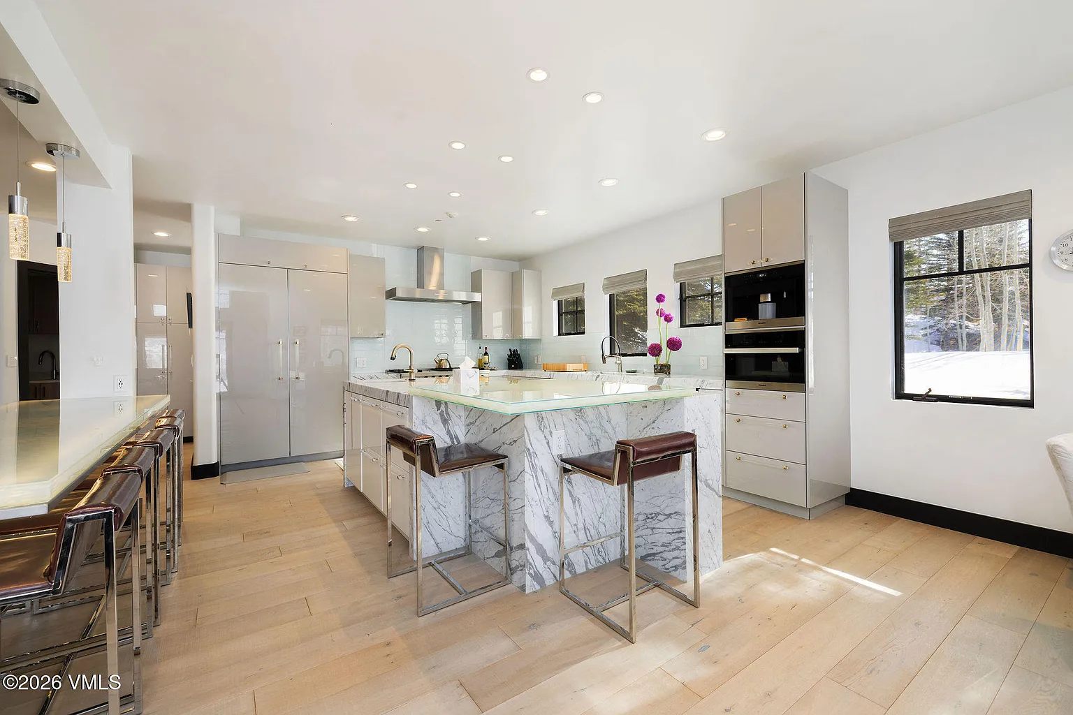 This is a bright, modern kitchen featuring glossy white cabinetry and countertops. The kitchen island is clad in marble and has a glass countertop extension, with stainless steel stools at the island. Light wood flooring adds warmth, and windows provide natural light.