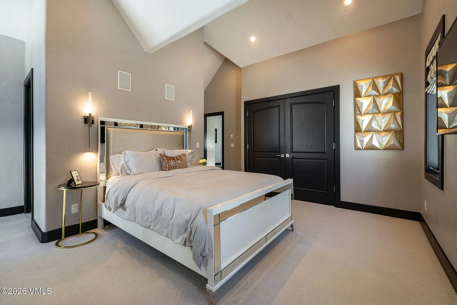 This primary bedroom showcases a sophisticated design with a neutral color palette. The room features a large mirrored bed frame, plush carpeting, and modern sconces. A unique geometric wall art piece and dark closet doors add visual interest, creating a serene and elegant atmosphere.