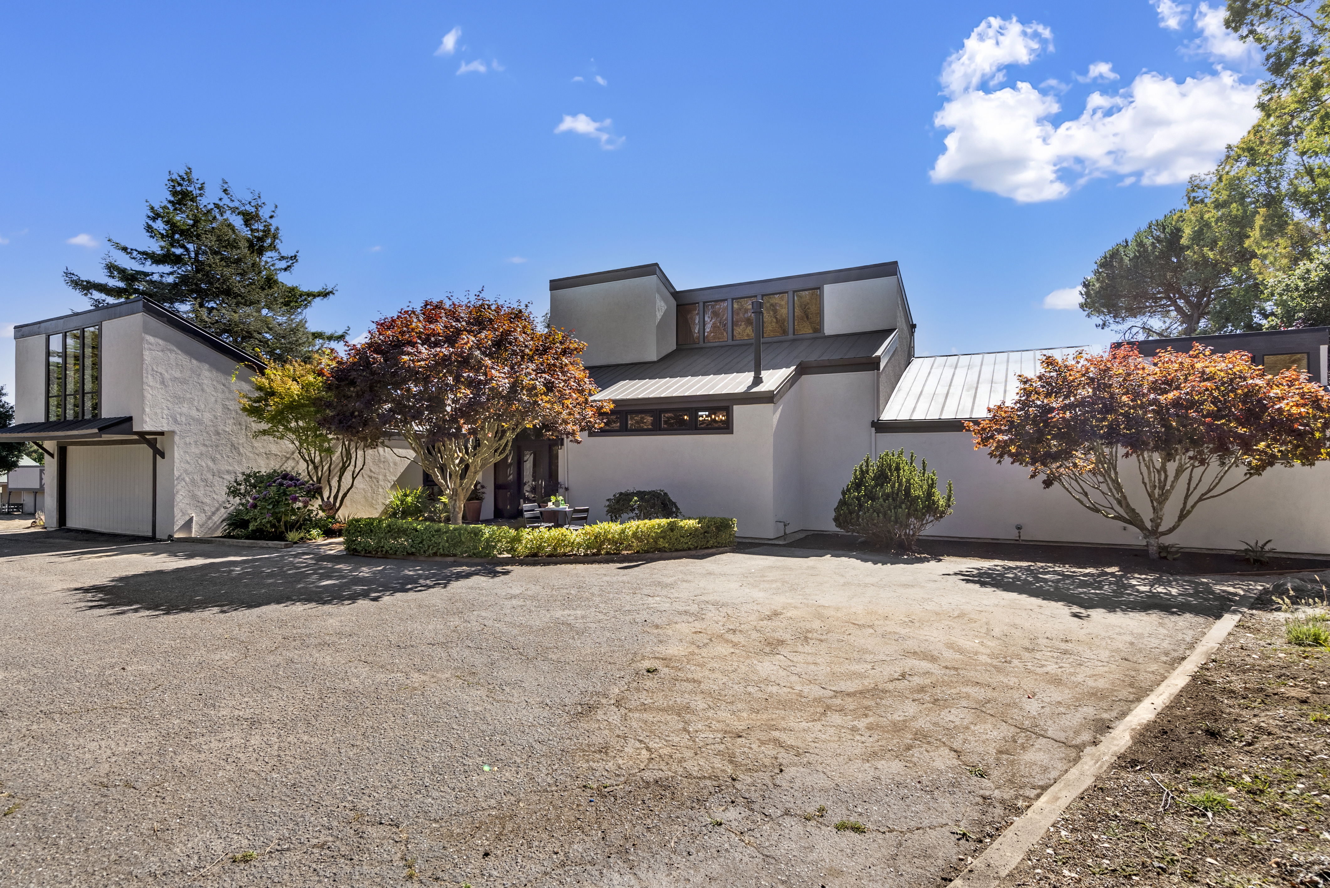 This modern contemporary home features a unique multi-level roofline and smooth stucco exterior, complemented by mature, vibrant Japanese maple trees in the front landscape. The property includes a paved driveway leading to an attached garage and a small patio area near the entrance, creating a welcoming and sophisticated curb appeal.