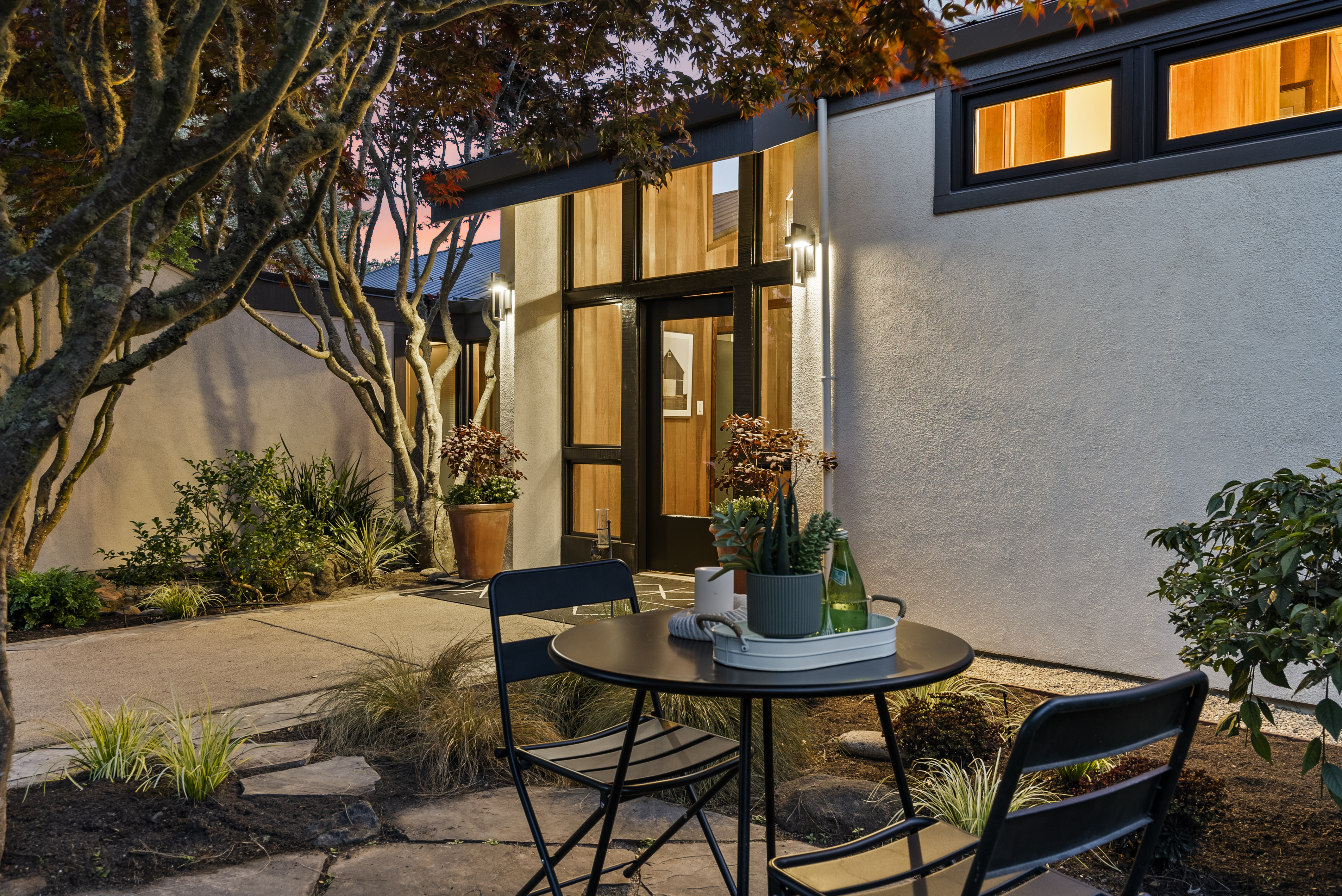 This charming modern entryway features a prominent glass-paneled door framed by floor-to-ceiling windows and warm wood paneling. The patio area is tastefully furnished with a bistro set, creating an inviting transition space nestled among lush, well-maintained landscaping and mature trees. The thoughtful exterior lighting and clean design lines evoke a sophisticated, contemporary aesthetic perfect for welcoming guests.
