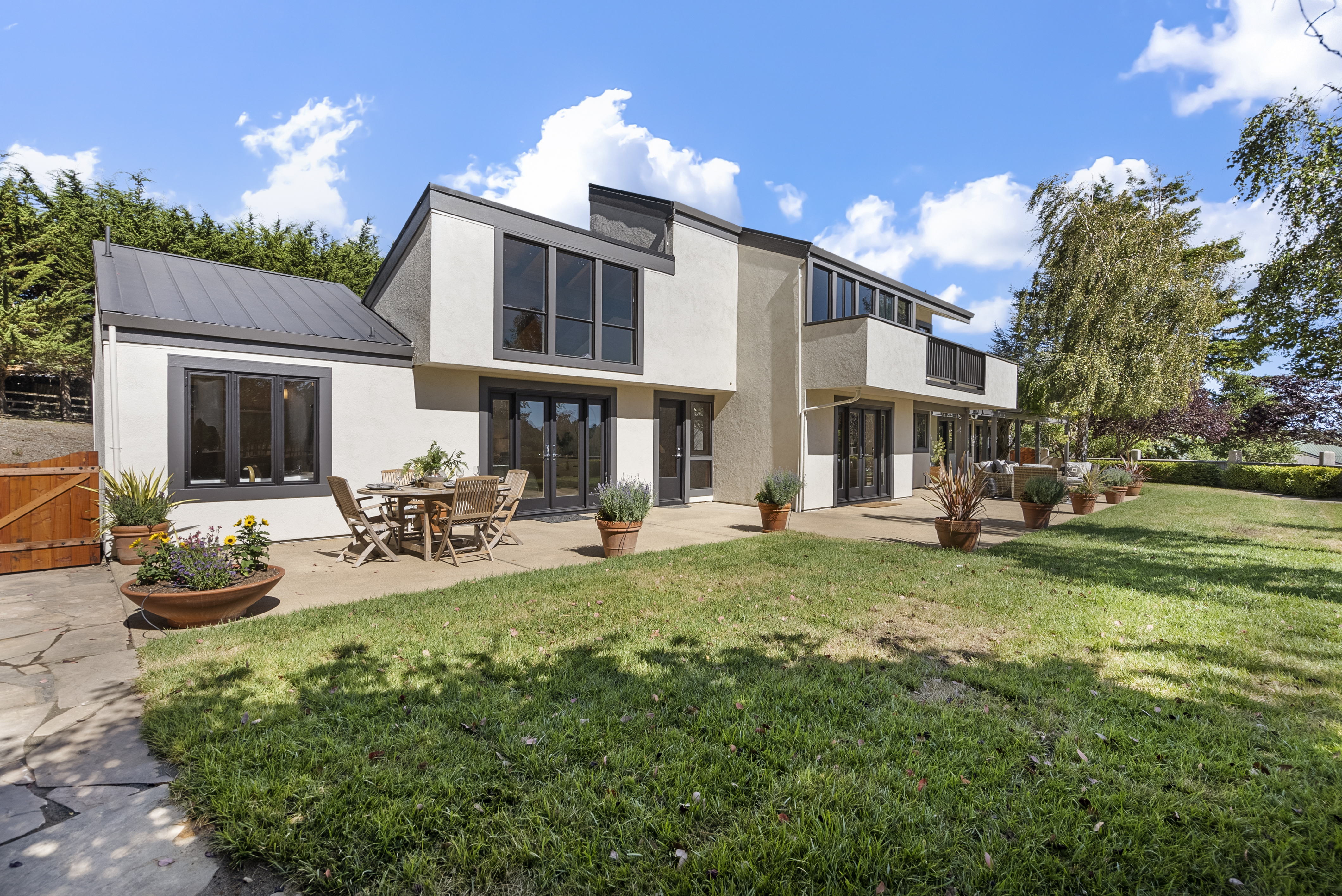 This modern home features a spacious rear elevation with a large, inviting concrete patio perfect for outdoor dining and entertaining. The exterior showcases a contemporary mixed-material design with clean lines, large windows, and a combination of soft-hued stucco and dark-framed accents. The property flows seamlessly into a well-maintained, grassy backyard, surrounded by established trees and lush landscaping, creating a serene and private living environment.