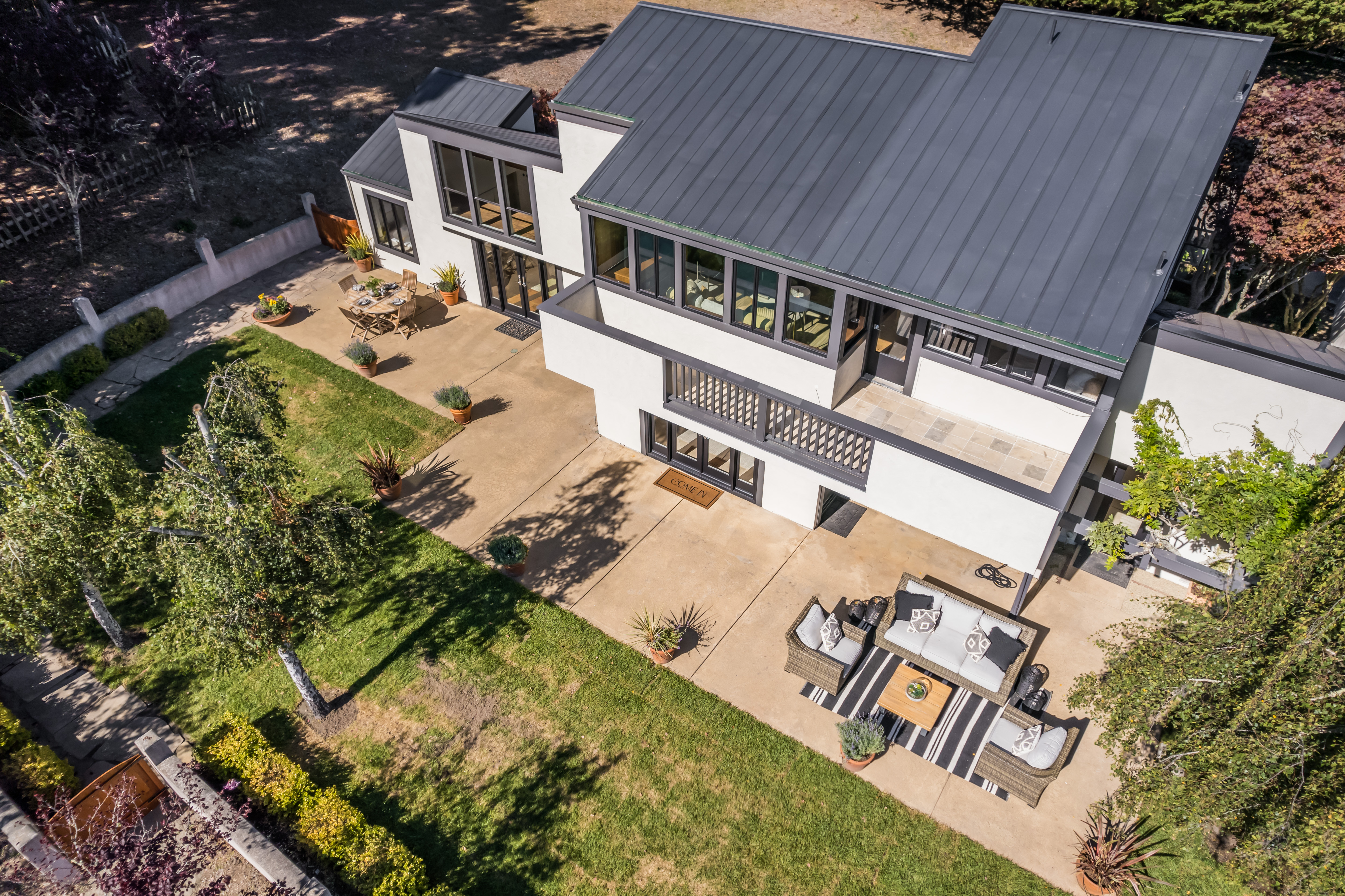 This high-angle aerial view showcases a modern, multi-level residence with a distinct dark metal roof and white stucco facade. The property features a spacious, shaded patio area set up for outdoor living and dining, transitioning smoothly into a well-maintained lawn. The aesthetic is contemporary and clean, emphasizing a seamless indoor-outdoor lifestyle perfect for entertaining.