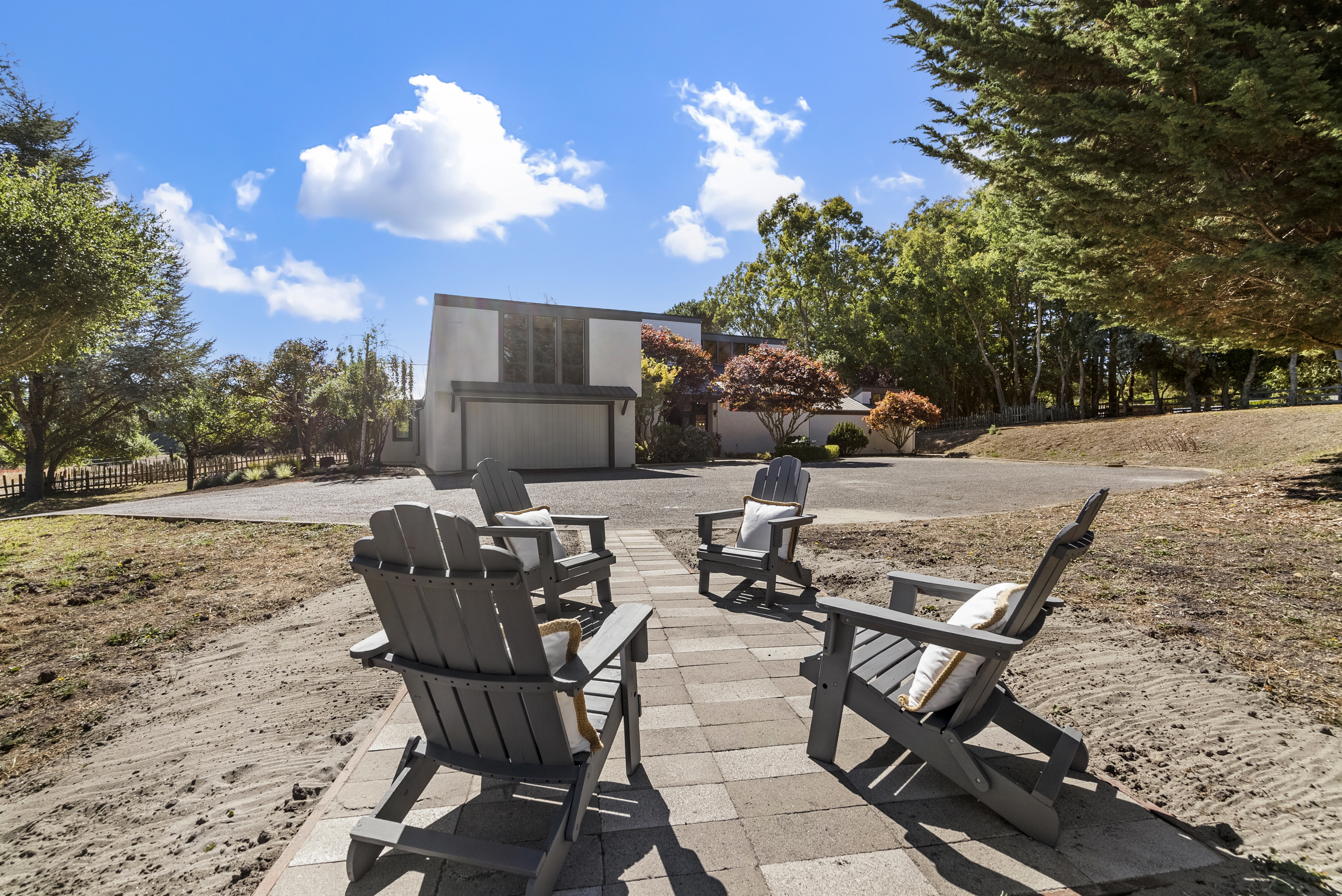 This outdoor space features a paved stone seating area arranged with four grey Adirondack chairs, ideal for relaxation or entertaining. The setting is surrounded by a natural, open landscape with mature trees, established lawn areas, and a view of a contemporary-style home in the background. The bright blue sky and sunny exposure create a welcoming and serene atmosphere.