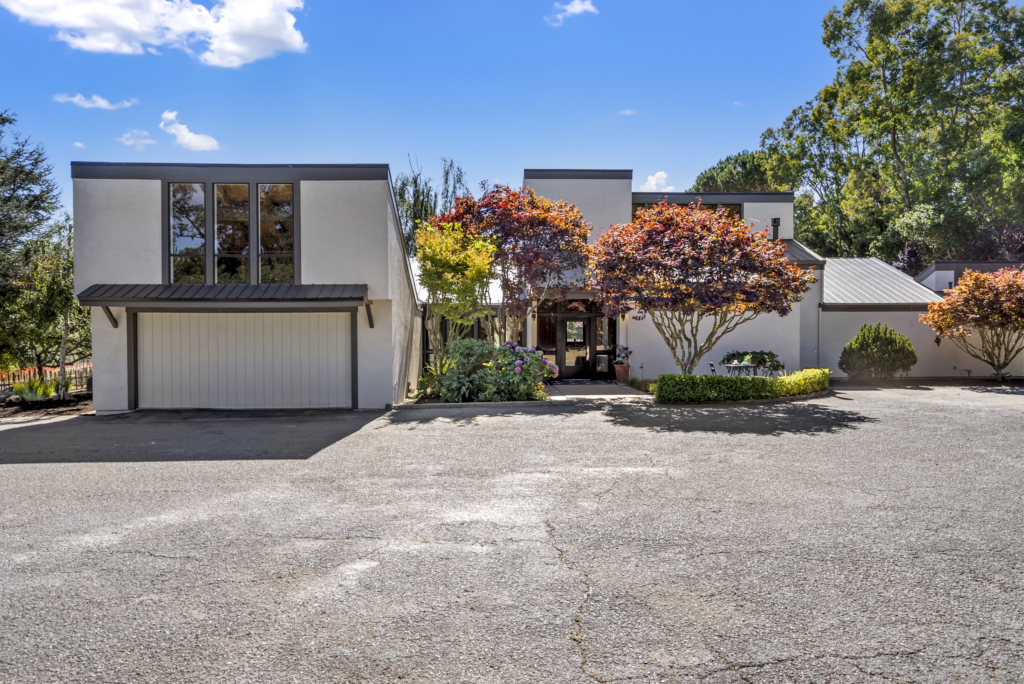This modern home features a distinct mid-century inspired architectural design with clean lines and a prominent two-car garage. The front exterior is accented by mature, vibrant trees with varied foliage and manicured landscaping, providing a welcoming and established curb appeal. A wide, paved driveway approaches the recessed front entryway, showcasing the property's serene and well-maintained setting.
