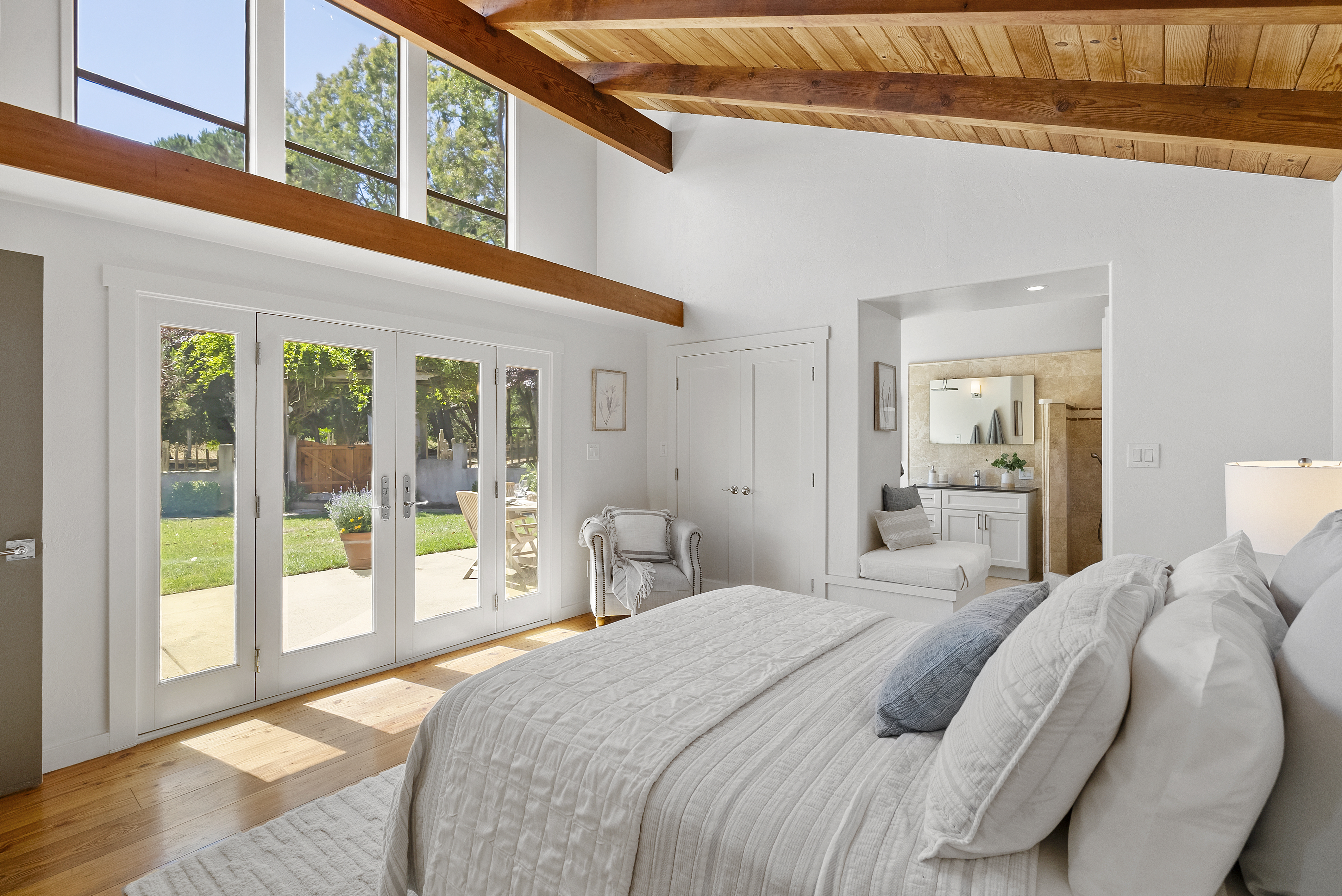 This spacious primary bedroom features a vaulted wood-beam ceiling, warm hardwood floors, and large glass double doors that open onto a sunny patio. The room includes a cozy seating nook with a comfortable armchair and a seamless transition into a private en-suite bathroom. Its bright, airy atmosphere combined with neutral textures creates a serene and inviting retreat.