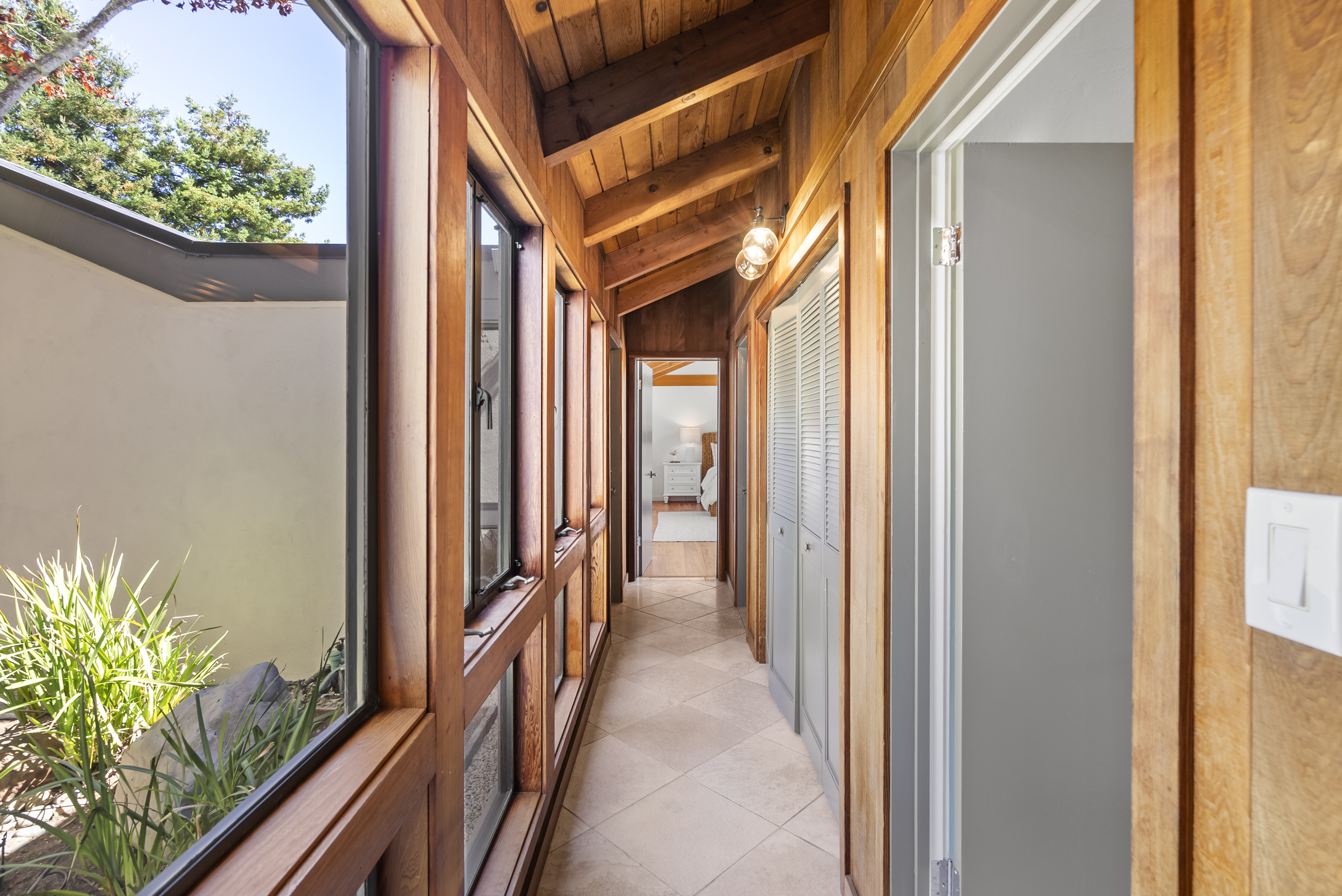This interior hallway features a distinctive rustic style with exposed wooden beams along the vaulted ceiling and natural wood wall paneling. Large floor-to-ceiling windows on the left frame a view of a tranquil interior garden, while the floor consists of light-toned tile arranged in a diagonal pattern. The design emphasizes natural light and creates a warm, connected transition between rooms.