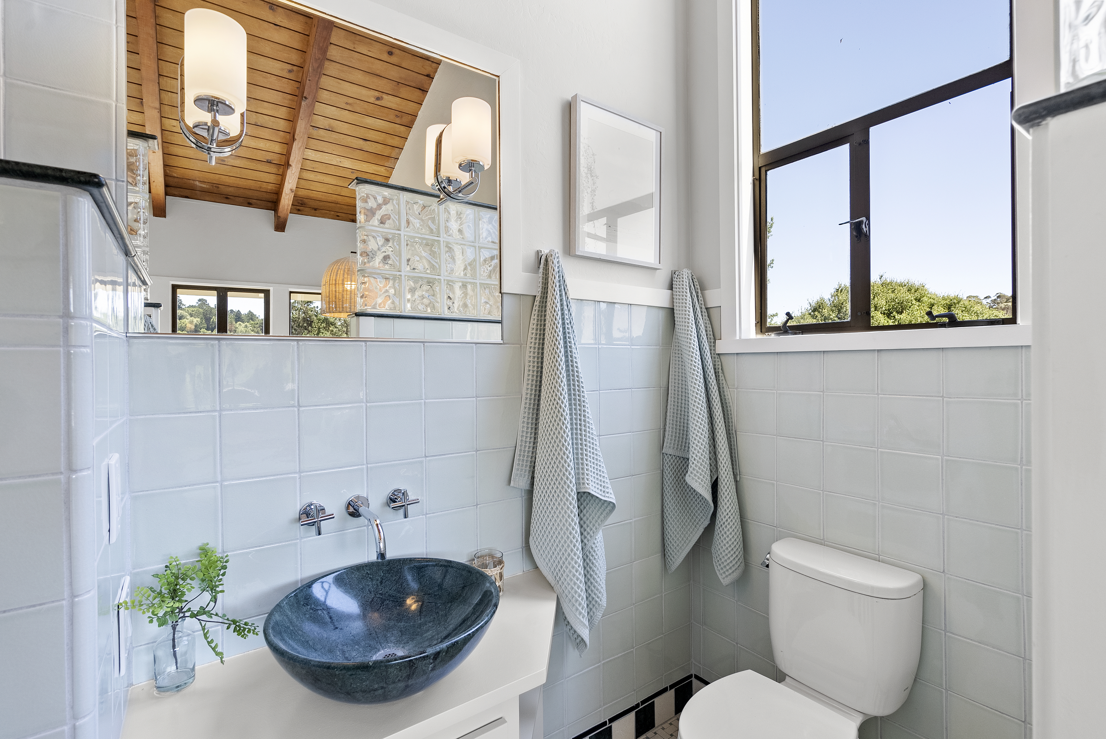 This bright and airy bathroom features a unique, dark stone basin sink set against light blue square wall tiles. A beautiful wood-paneled, vaulted ceiling is reflected in the wall mirror, adding a touch of rustic warmth, while a large window lets in plenty of natural light. Waffle-weave towels and minimalist fixtures complete the coastal, clean aesthetic of this space.