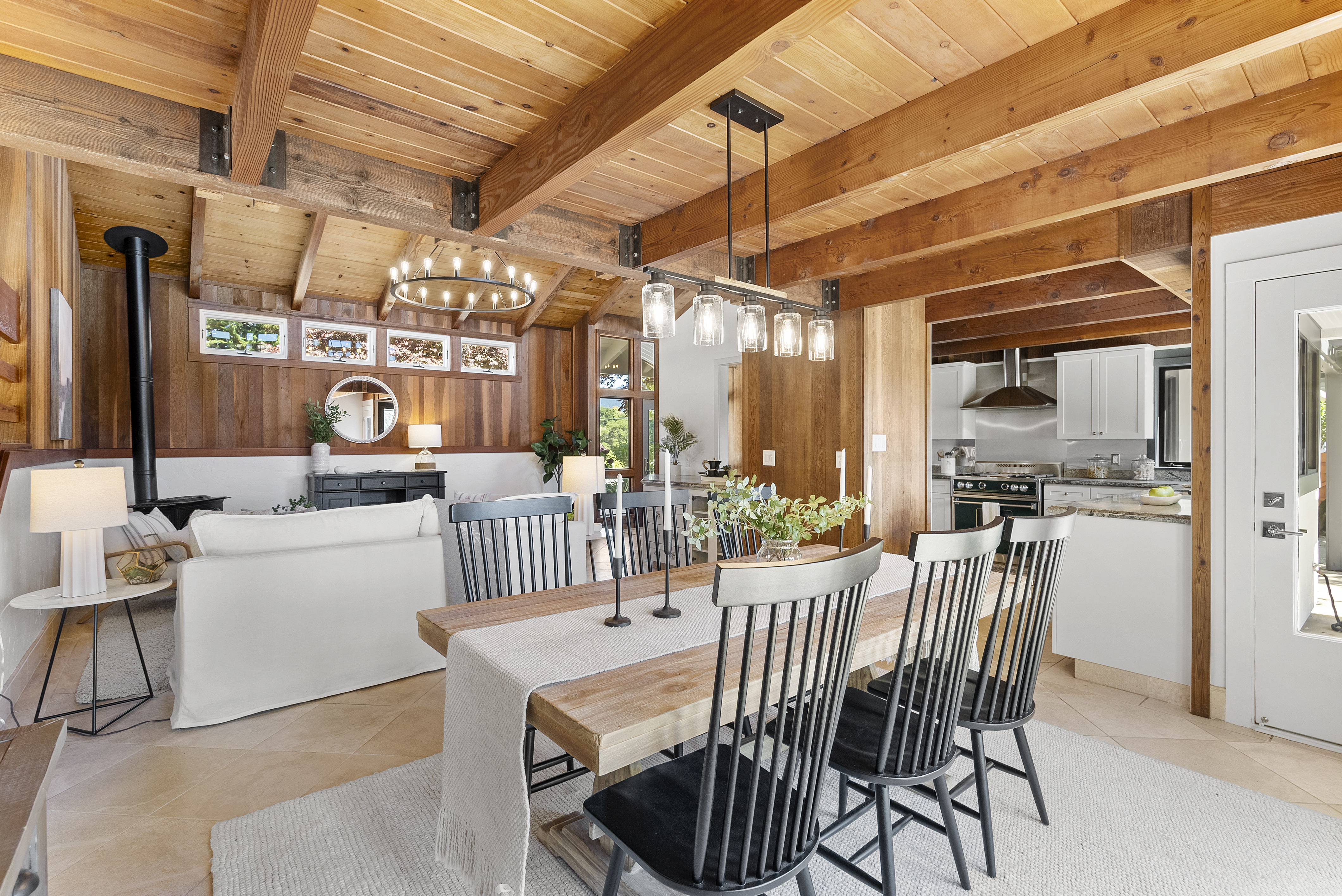 This bright, open-concept dining room exudes a warm, rustic-modern charm with its exposed natural wood ceiling beams and wood-paneled walls. The space features a heavy wooden dining table with spindle-back chairs, set under a stylish industrial-style pendant light, and flows seamlessly into a cozy living area and a bright white kitchen, making it ideal for entertaining. Large windows and light-colored tile flooring contribute to an airy and inviting atmosphere.