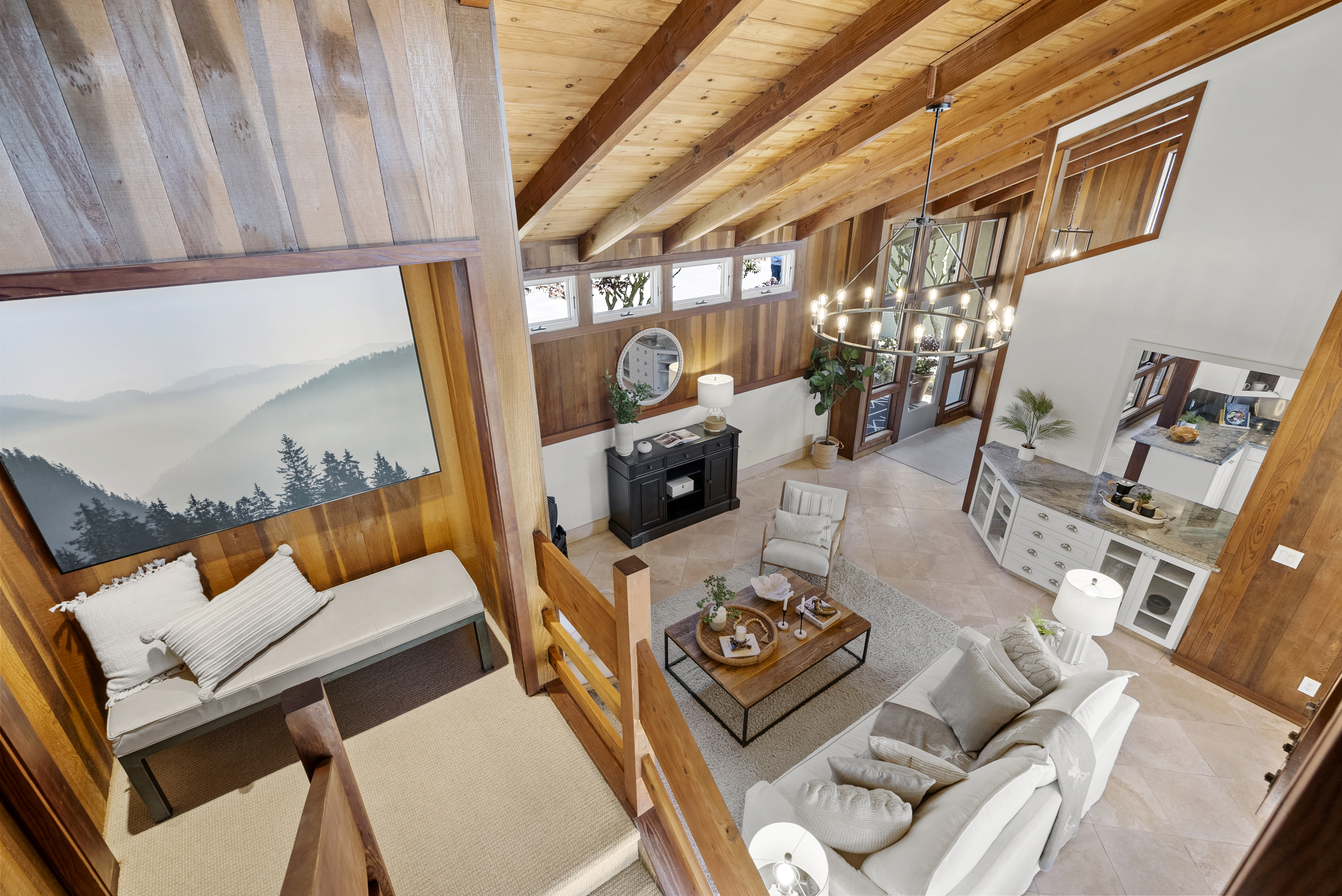 This living room features a striking vaulted ceiling with exposed wood beams and a large, artistic landscape mural above a cozy bench seat. The open, airy floor plan connects the seating area, anchored by a rustic coffee table and plush sofa, to a sleek adjacent kitchen with stone countertops. Natural light floods the space through clerestory windows, creating a warm and sophisticated mountain-lodge aesthetic.